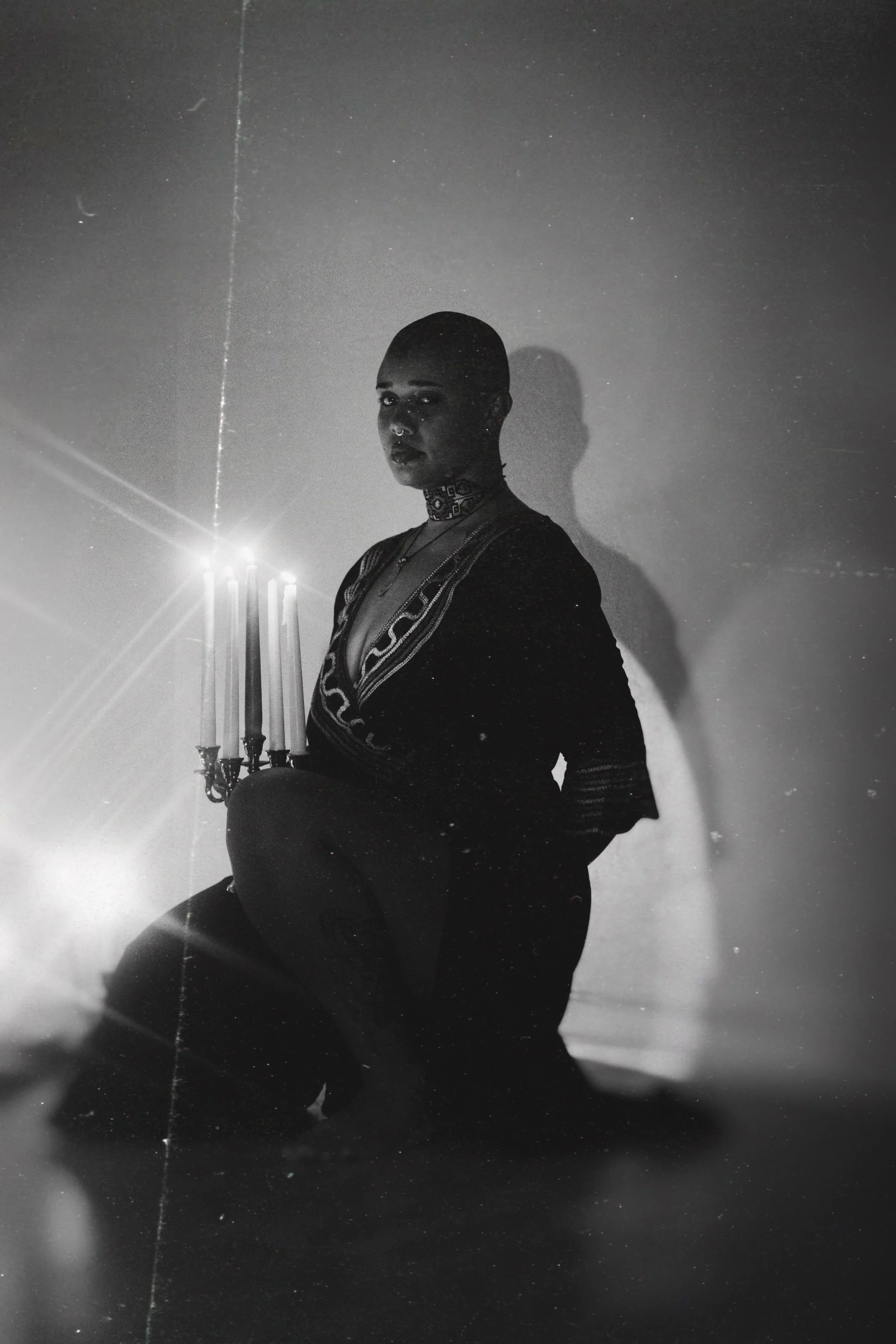 Black and white photo of a person with a shaved head, sitting on the floor, holding a tall candelabra with three lit candles, casting a shadow on the wall behind. The person is wearing a dark robe with intricate patterns, a nose ring, and a choker necklace.