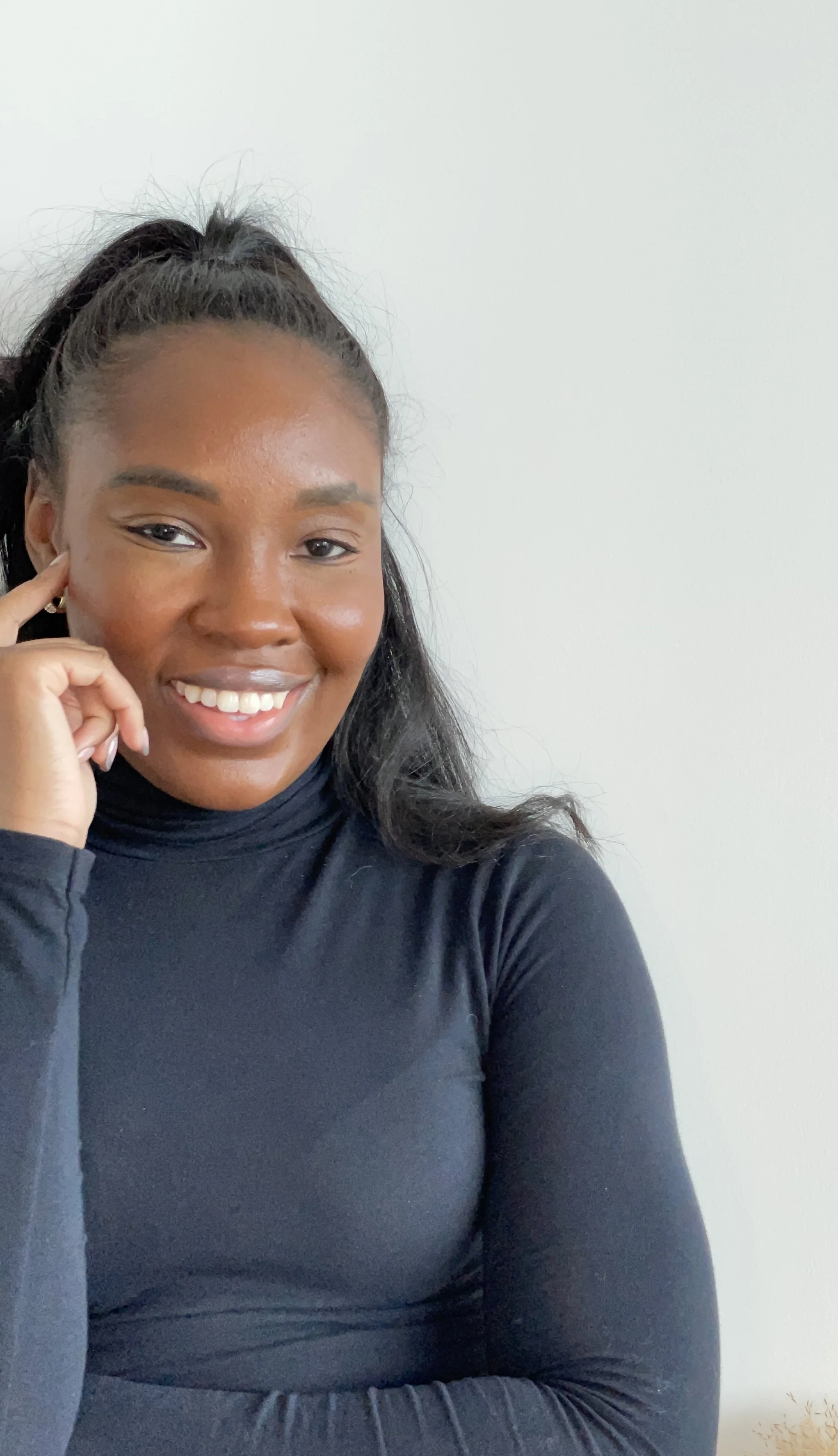 A young woman with dark hair styled in a ponytail, smiling and resting her chin on her hand, wearing a black turtleneck top against a plain light-colored background.