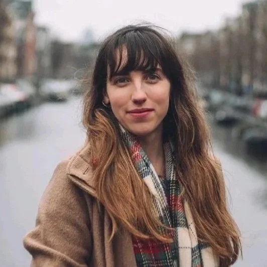 A young woman with long brown hair, wearing a tan coat and plaid scarf, standing in front of a canal with boats and European-style buildings.