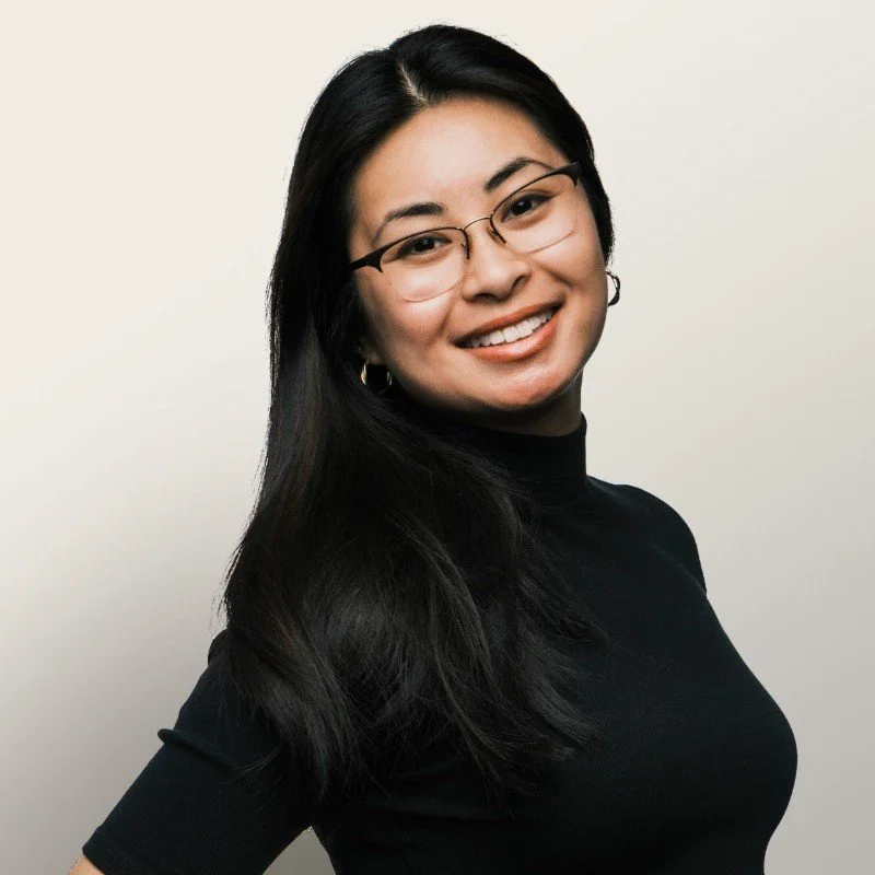 A woman with long black hair, glasses, and hoop earrings, smiling against a plain light-colored background.