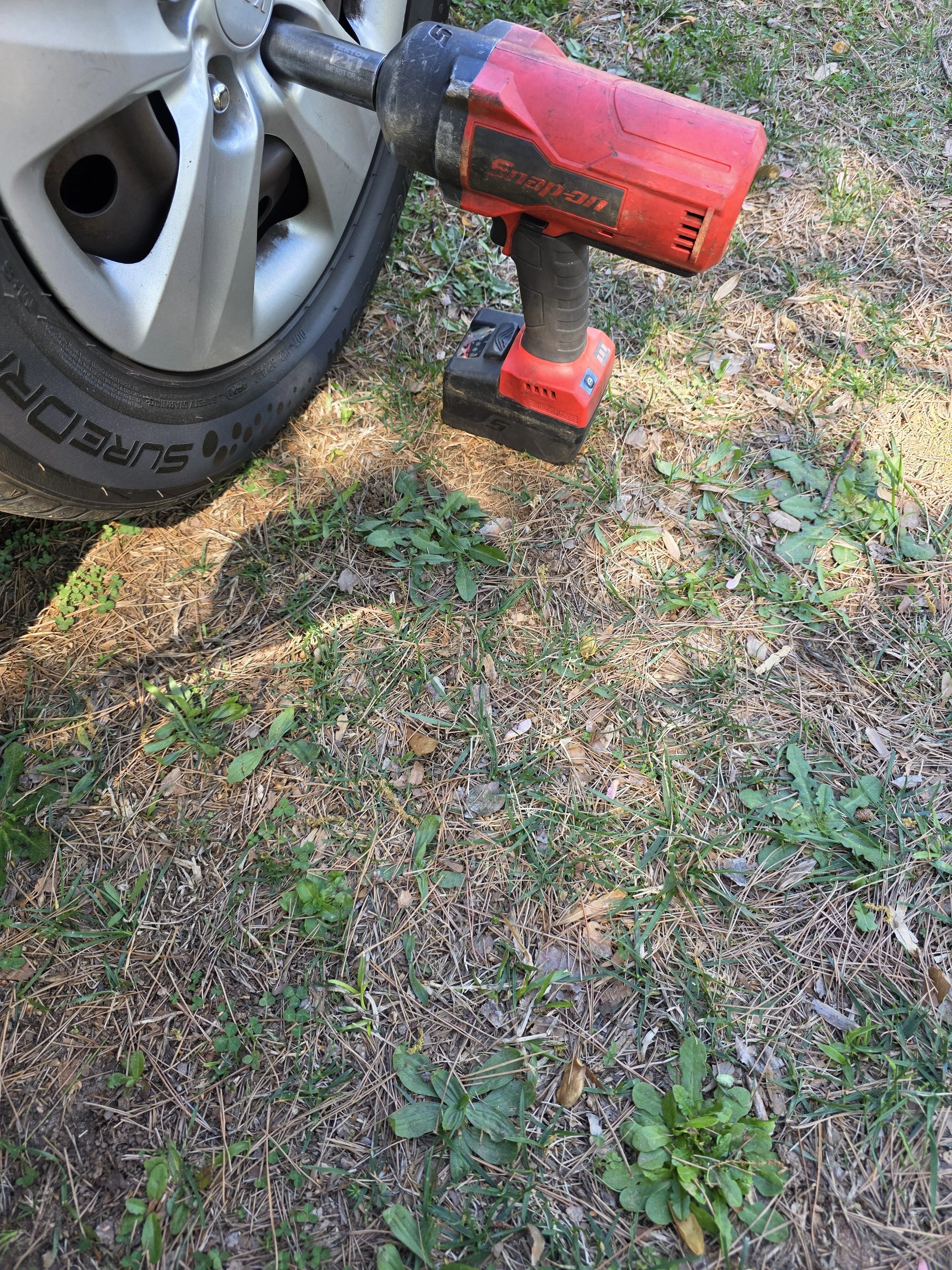 A red cordless drill resting against a car tire on a grassy ground with pine needles and small plants.