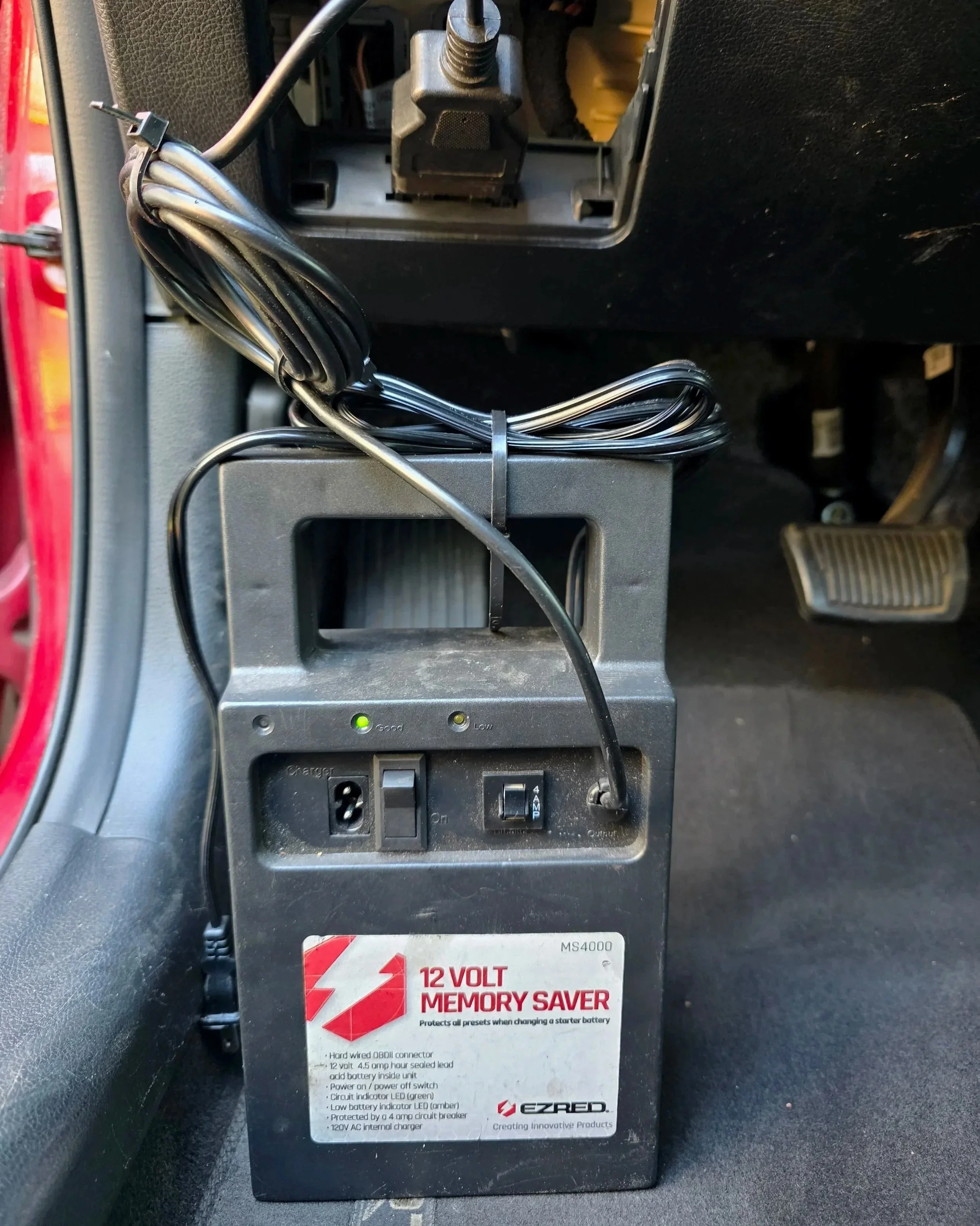 A 12-volt memory saver device on the floor of a vehicle, connected to the vehicle's electrical system with multiple wires, with indicator lights and switches on the device.