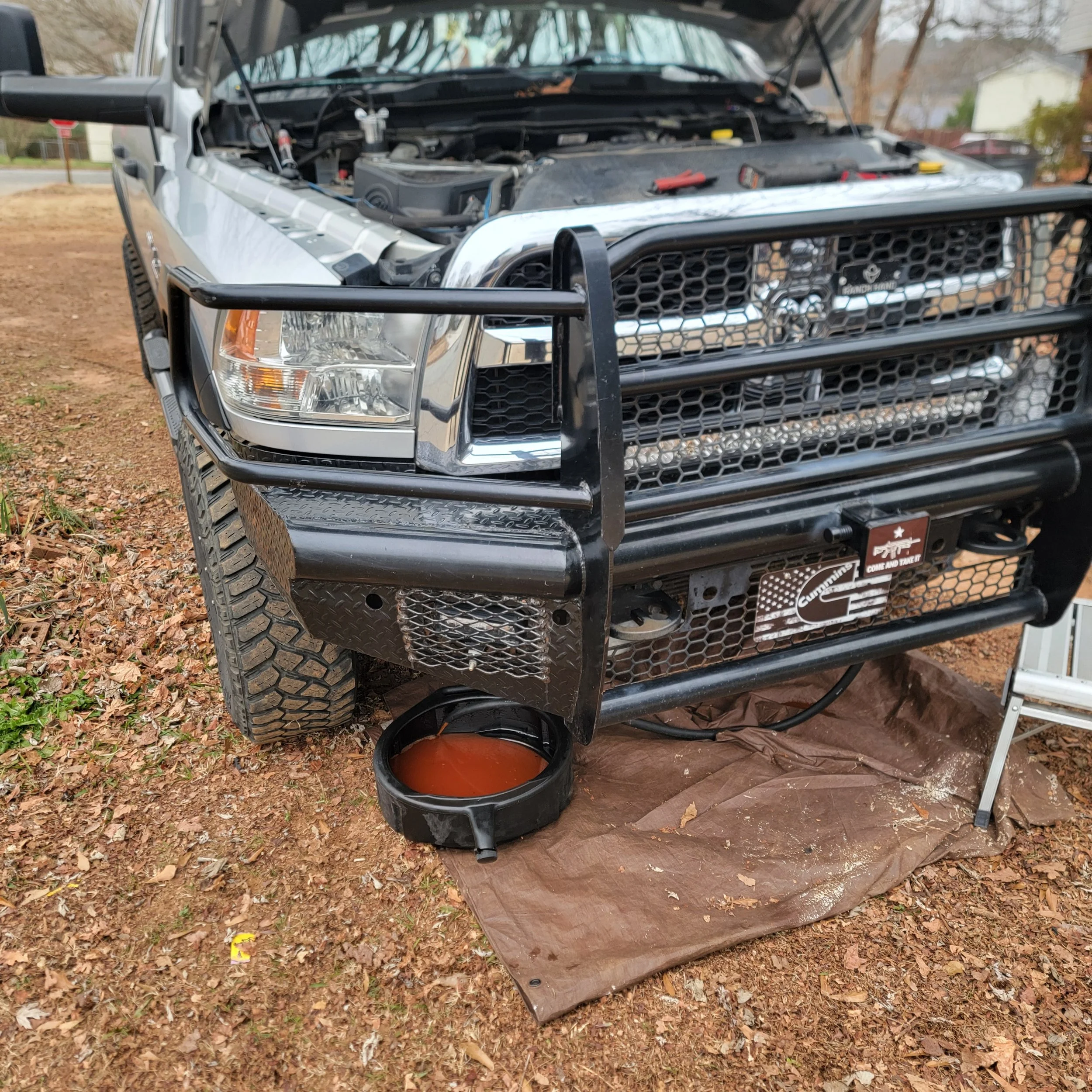 Front of a pickup truck with open hood, black grille guard, and an oil drain pan underneath collecting oil on a brown tarp on a dirt ground.