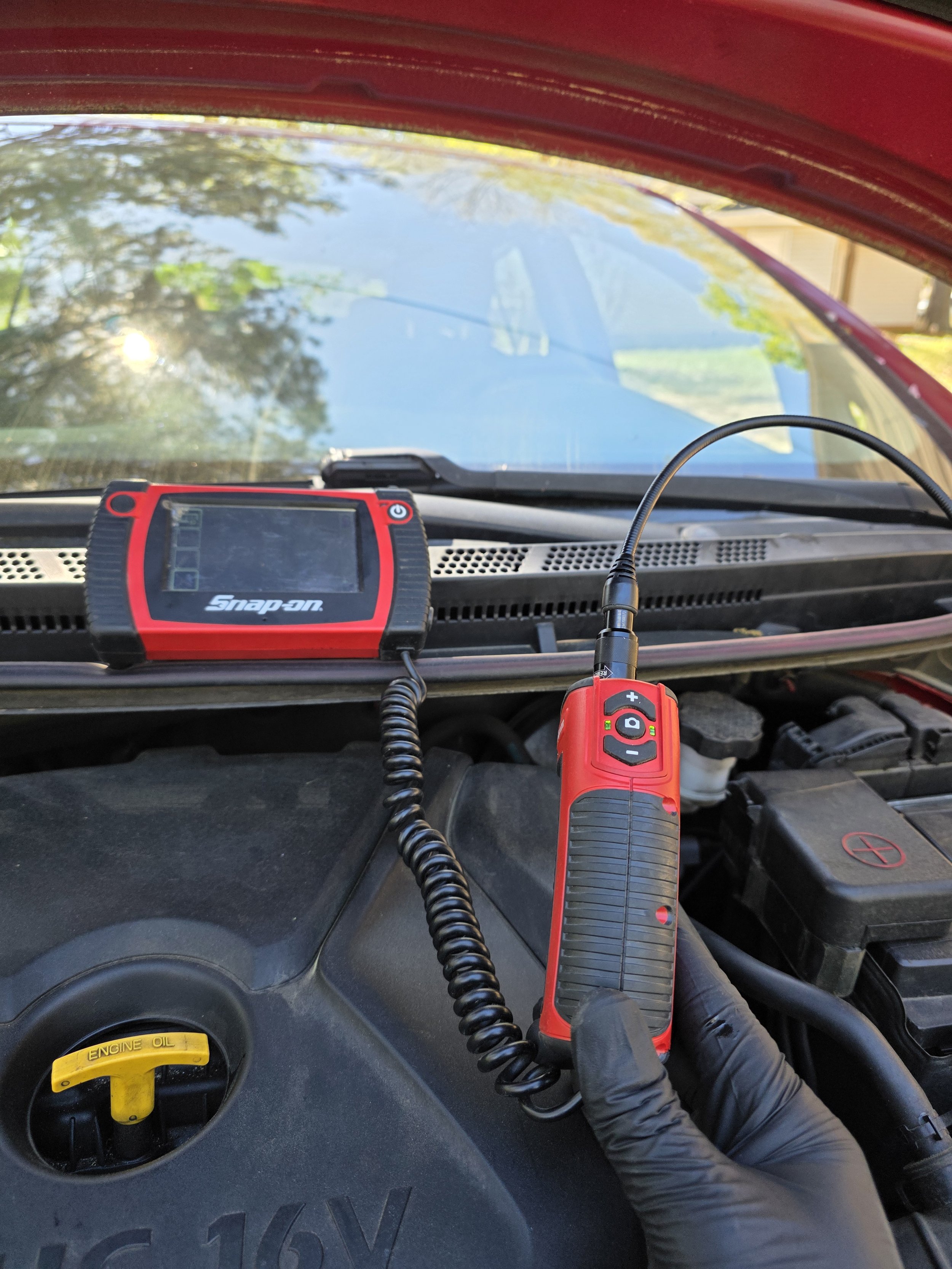 Inside a vehicle engine bay showing diagnostic tools, including a hand holding a red electronic device connected to a scanning tool on the dashboard, with the windshield and trees reflected on the glass.