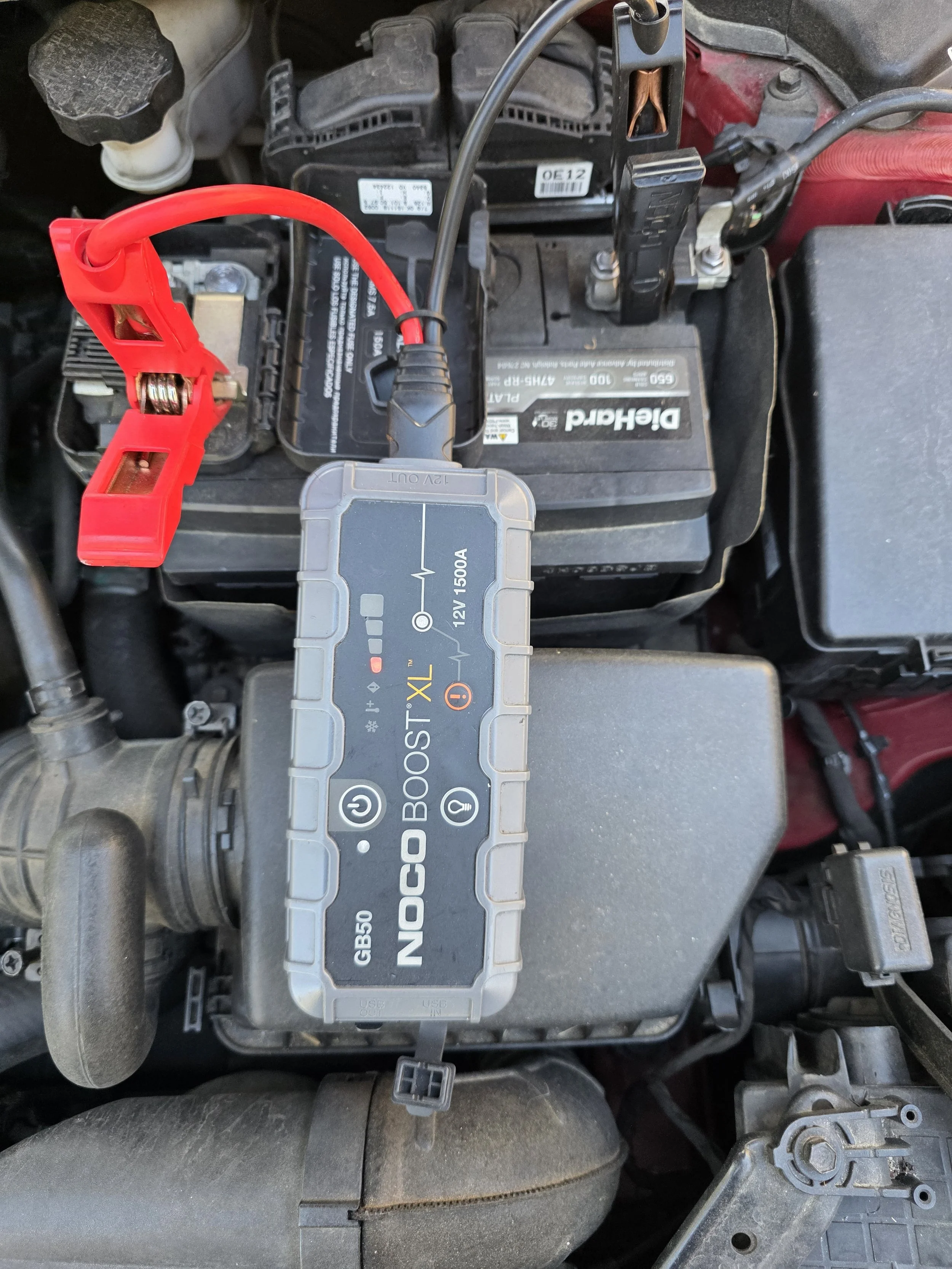A car battery with a Jump Boost XL device connected to the terminals under the hood of a vehicle.