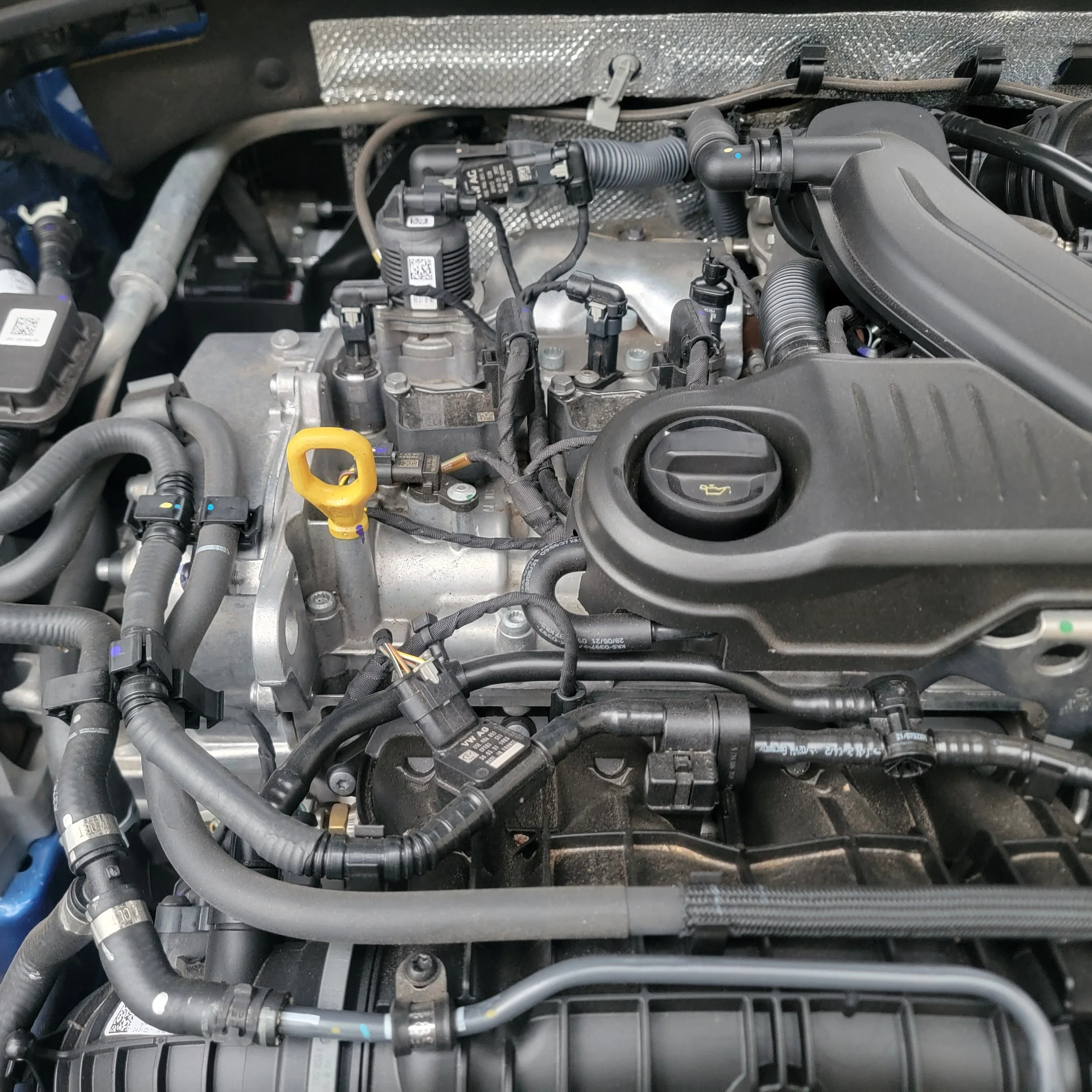 Close-up of a car engine bay showing various engine components, hoses, and wiring with a black engine cover and yellow oil dipstick.
