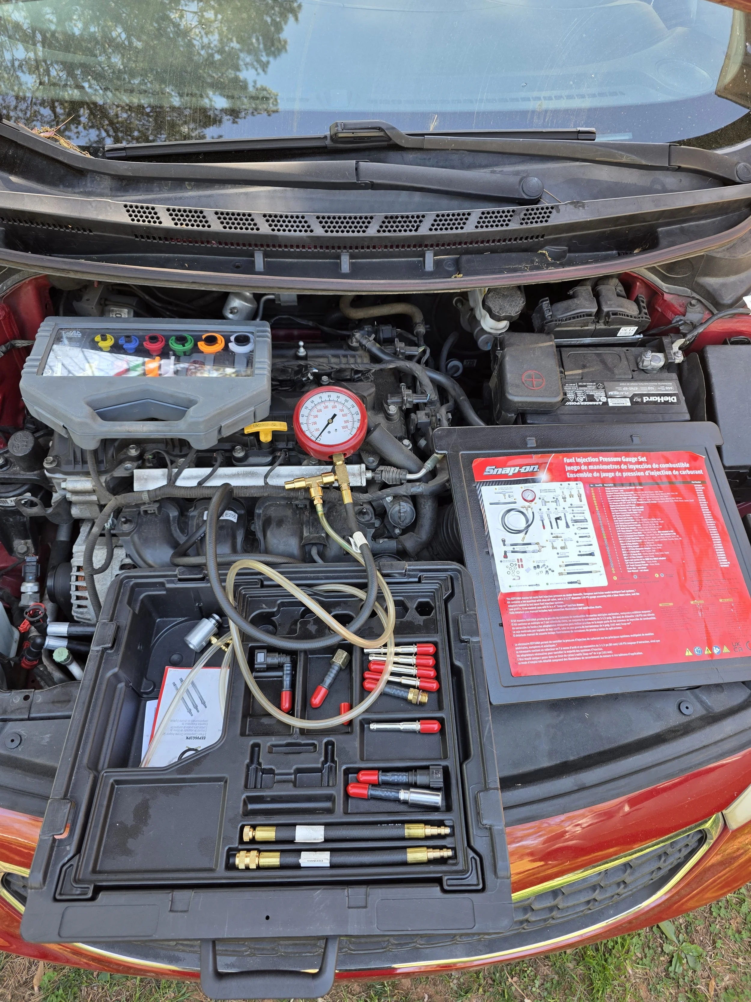 Car hood open, tools and test equipment on front of engine bay, with test gauge and hoses connected.