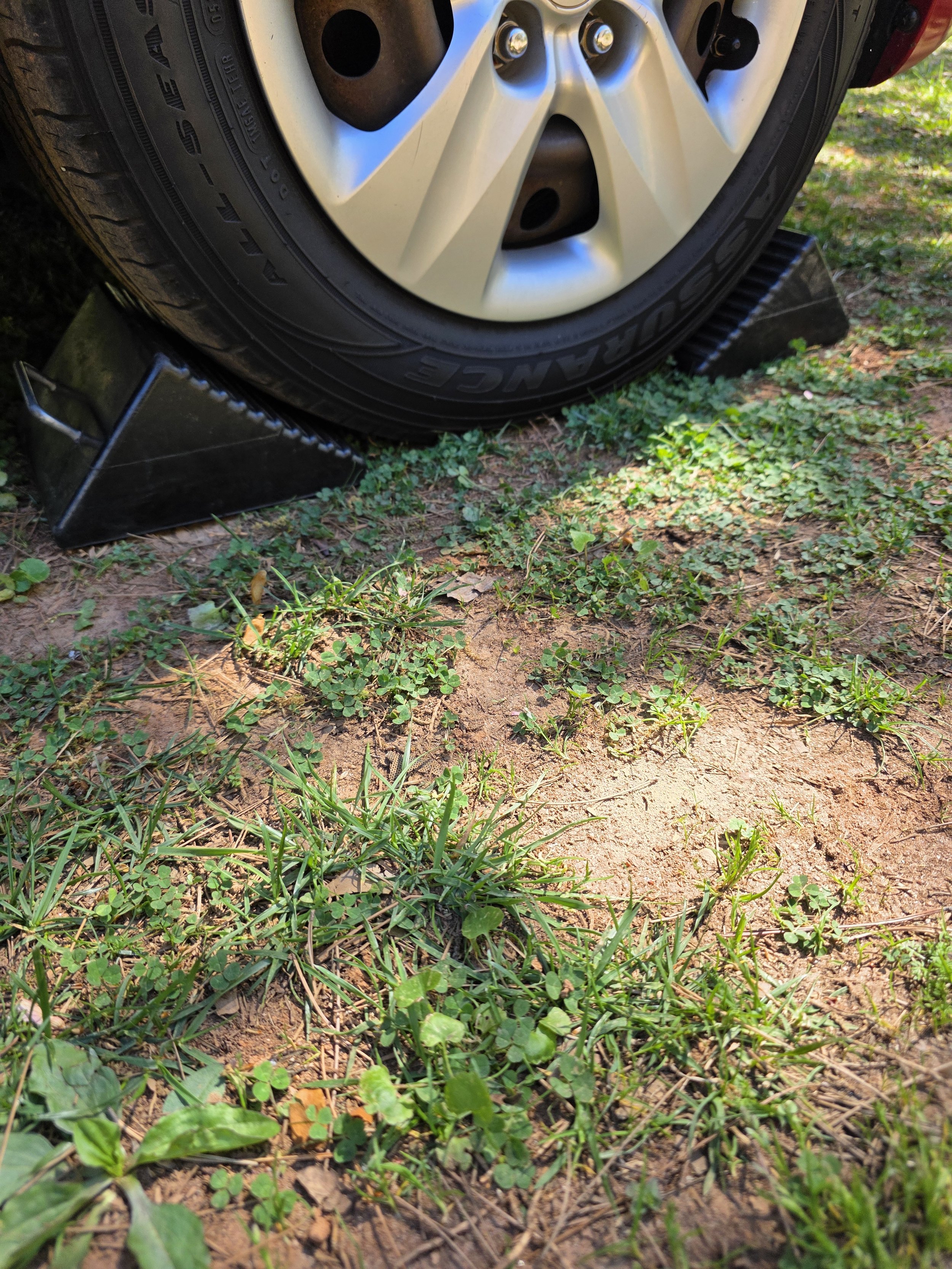 A car tire on a physical wheel chock, placed on dirt with grass and small plants growing through the soil.