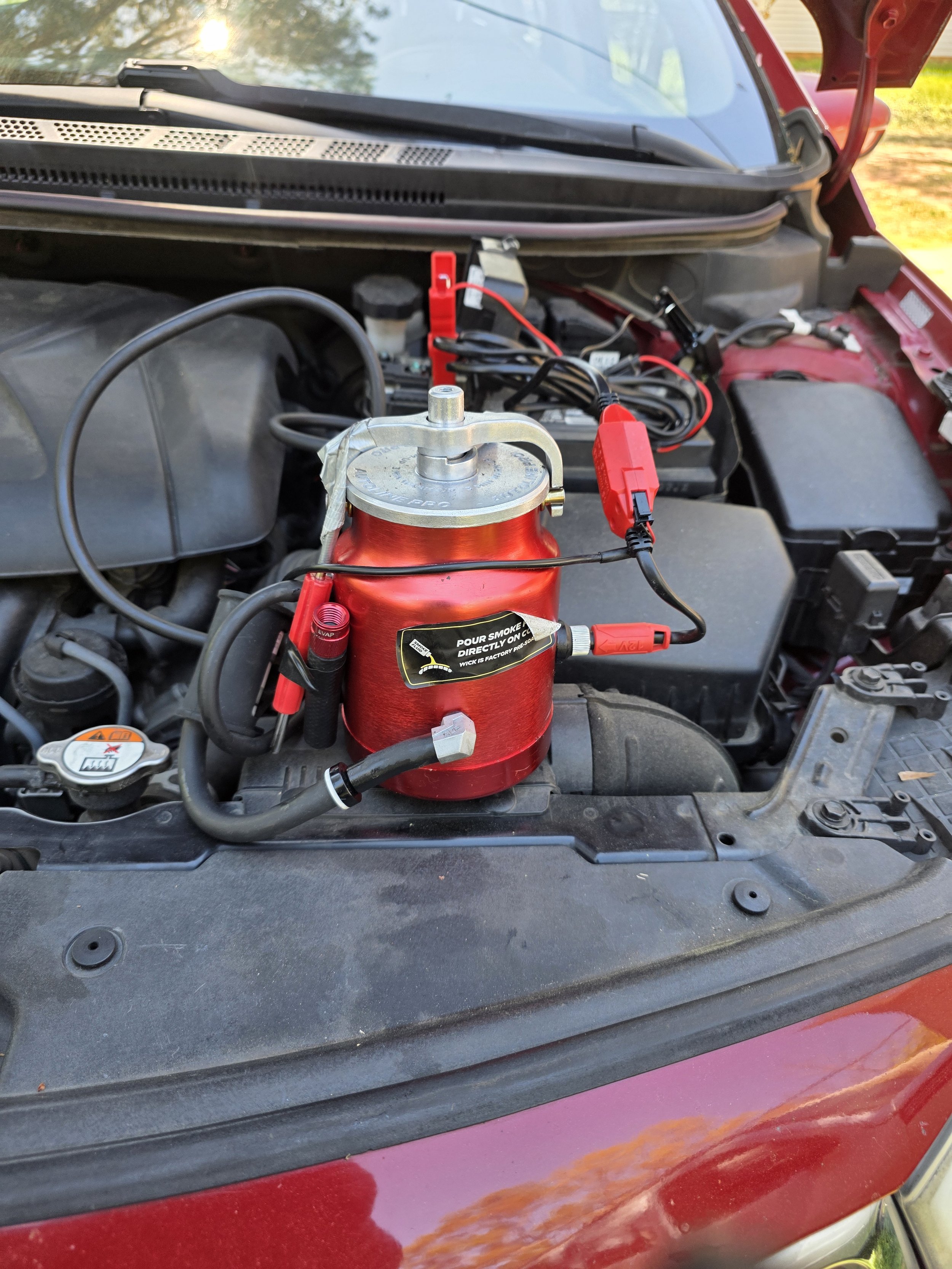 Car engine bay with a red portable jump starter attached to the battery terminals.