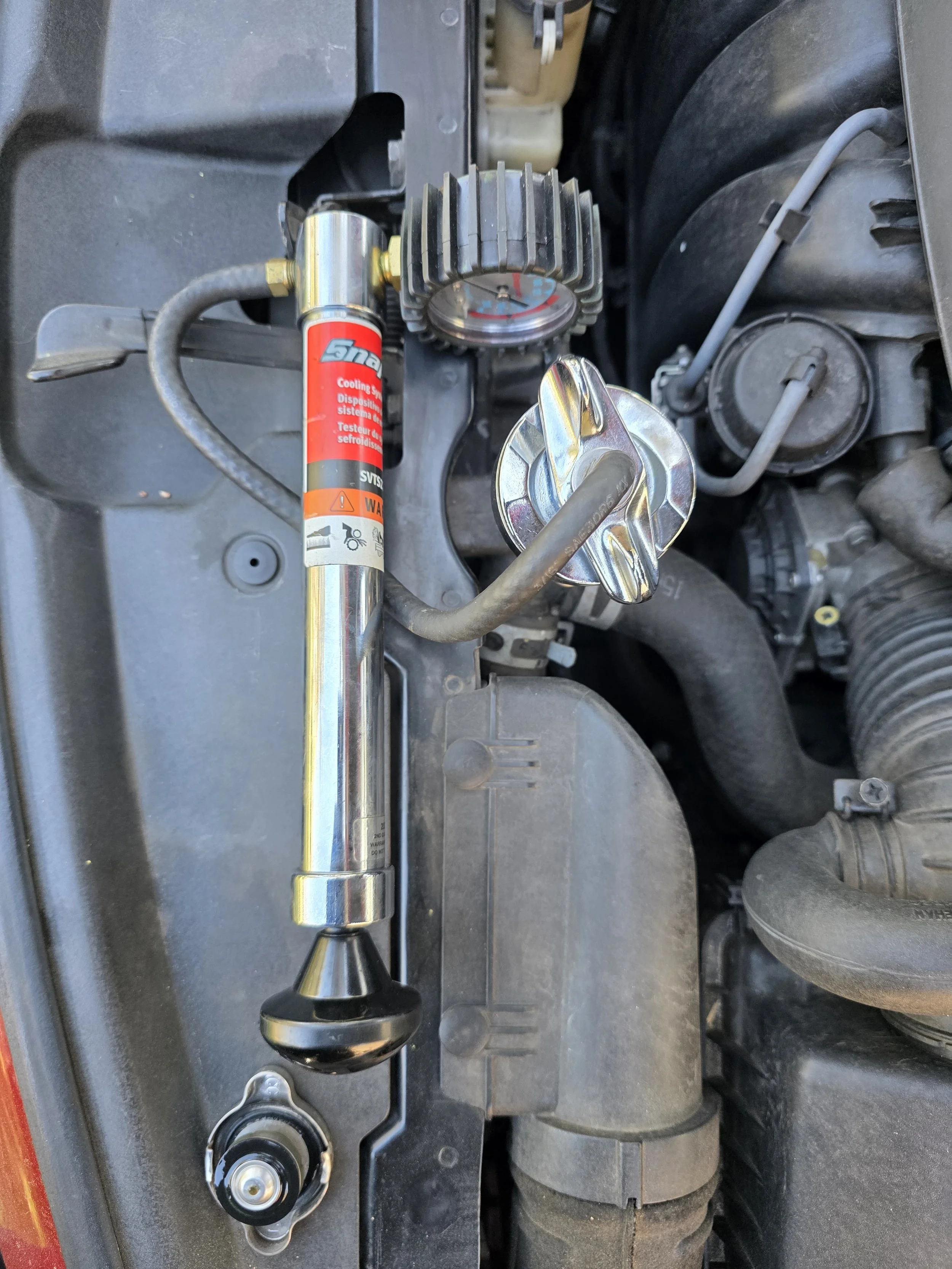 Under the hood of a vehicle, a shock absorber with a sticker reading 'Cooling System' is visible alongside a shiny metal cap, an engine component, and various hoses and pipes.