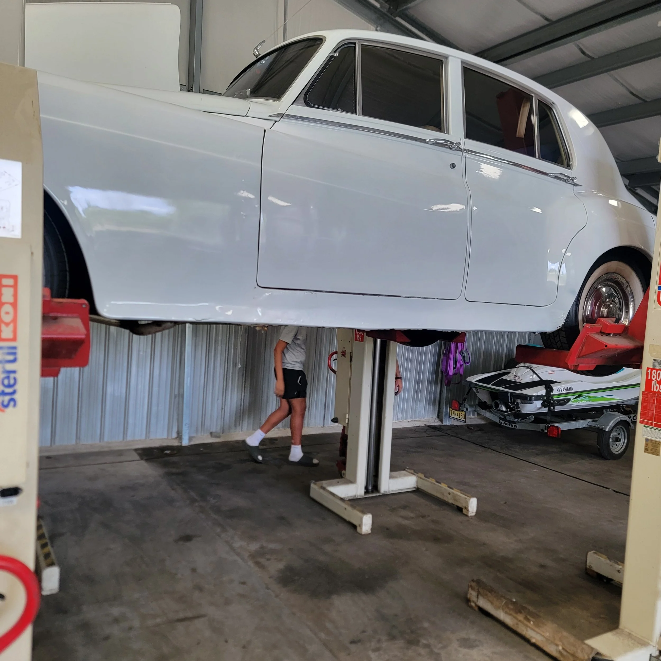 A vintage white car elevated on a hydraulic lift inside a garage, with a person standing underneath, partially visible. There is a small boat on a trailer in the background.