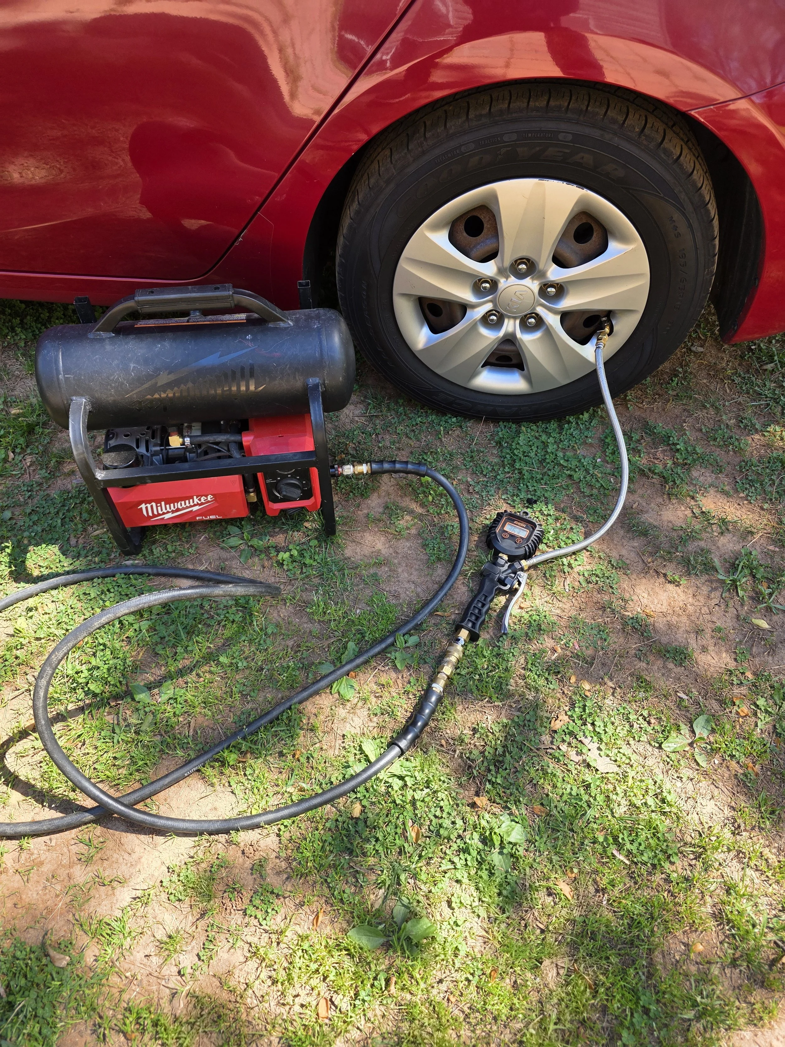 A red car with a tire connected to a portable air compressor via a hose, on a grassy patch.