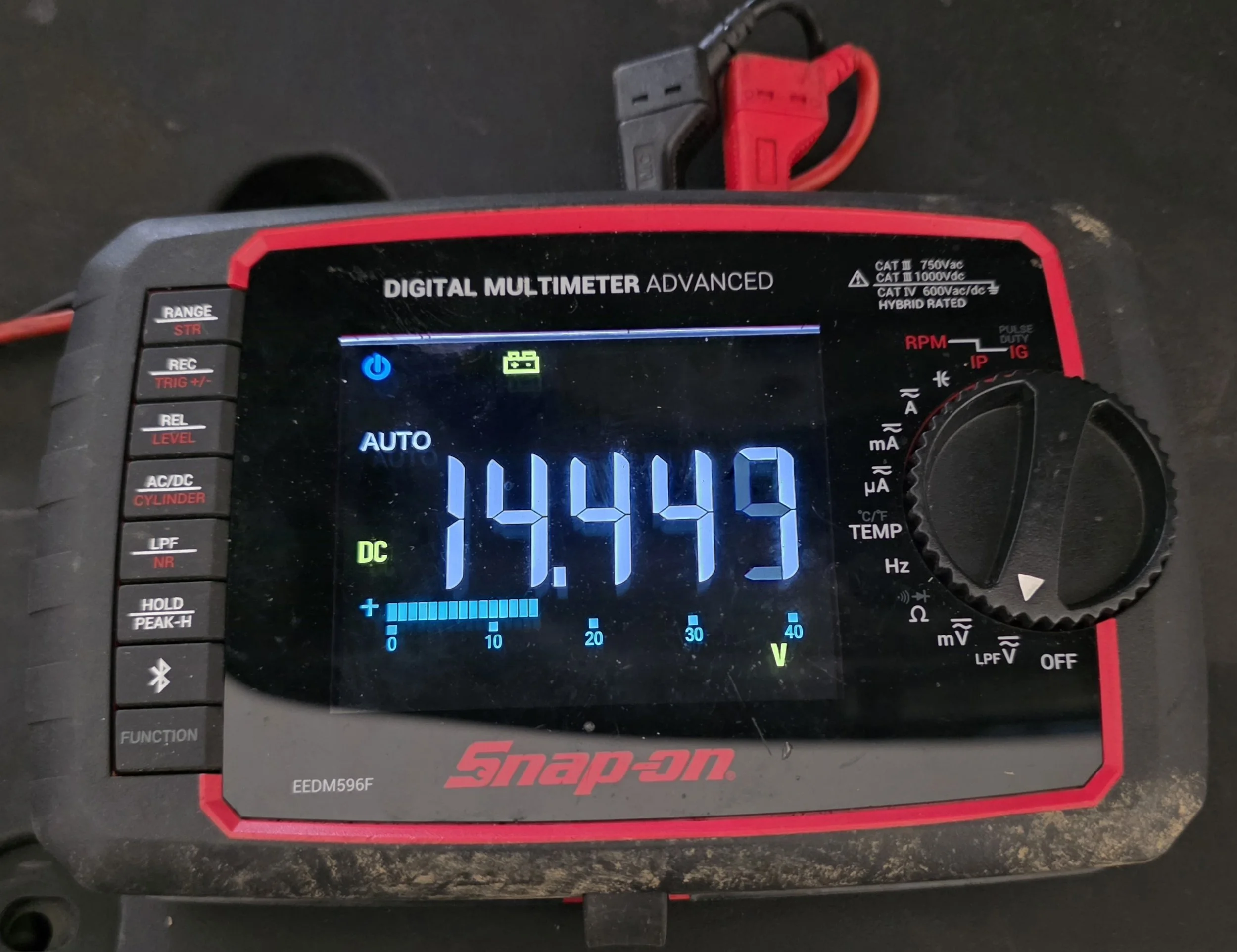 A digital multimeter displaying a voltage of 14.449 volts with a blue and black display. The multimeter is a Snap-on brand with various buttons on the left side and a rotary dial on the right. There is a small battery icon and other measurement indic