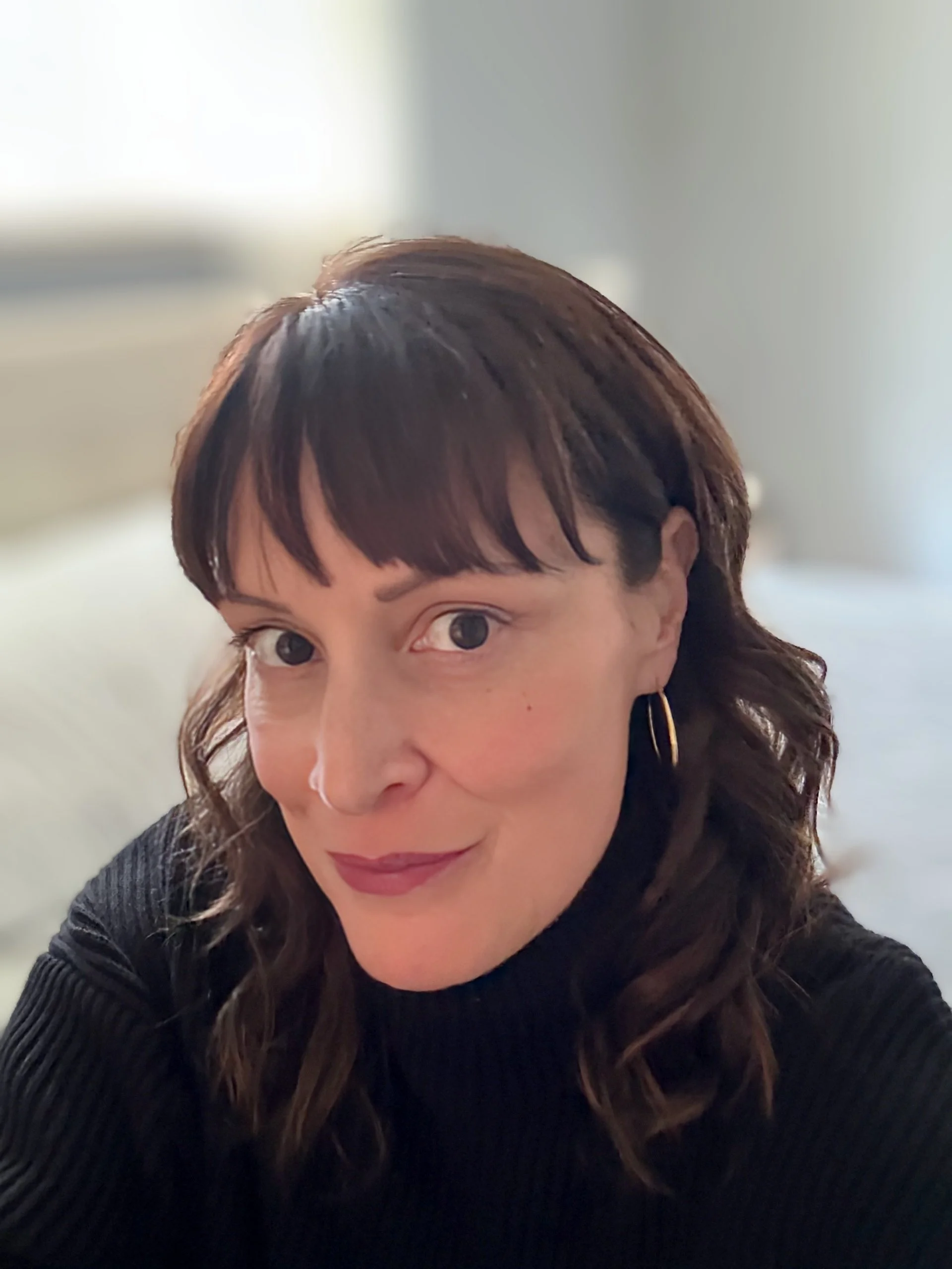 A woman with short dark brown hair, wearing hoop earrings and a black top, looking into the camera with a slight smile in a softly lit room.
