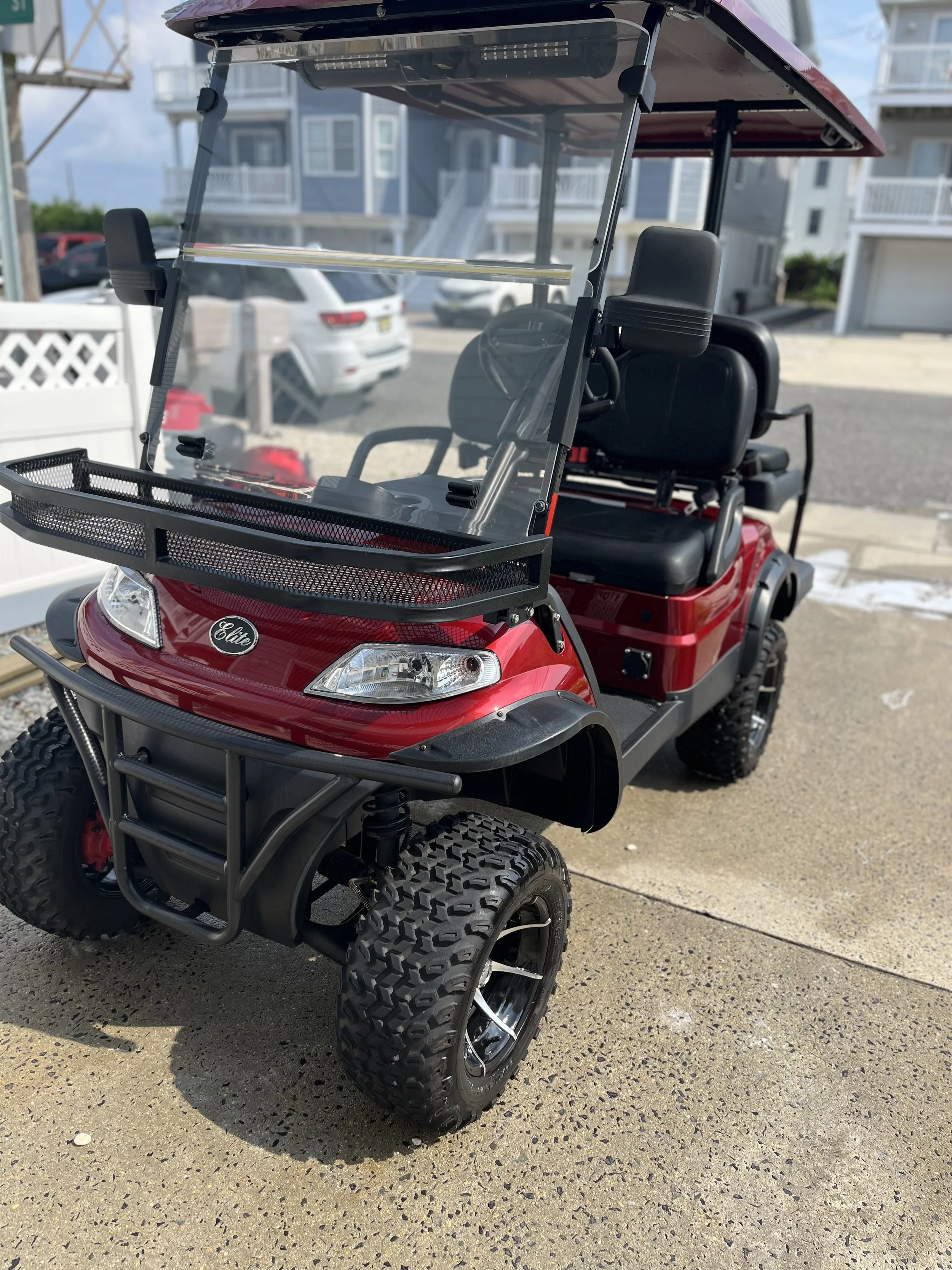 Red golf cart with black tires and windshield, parked on a paved surface outside residential buildings, with a clear sky overhead.