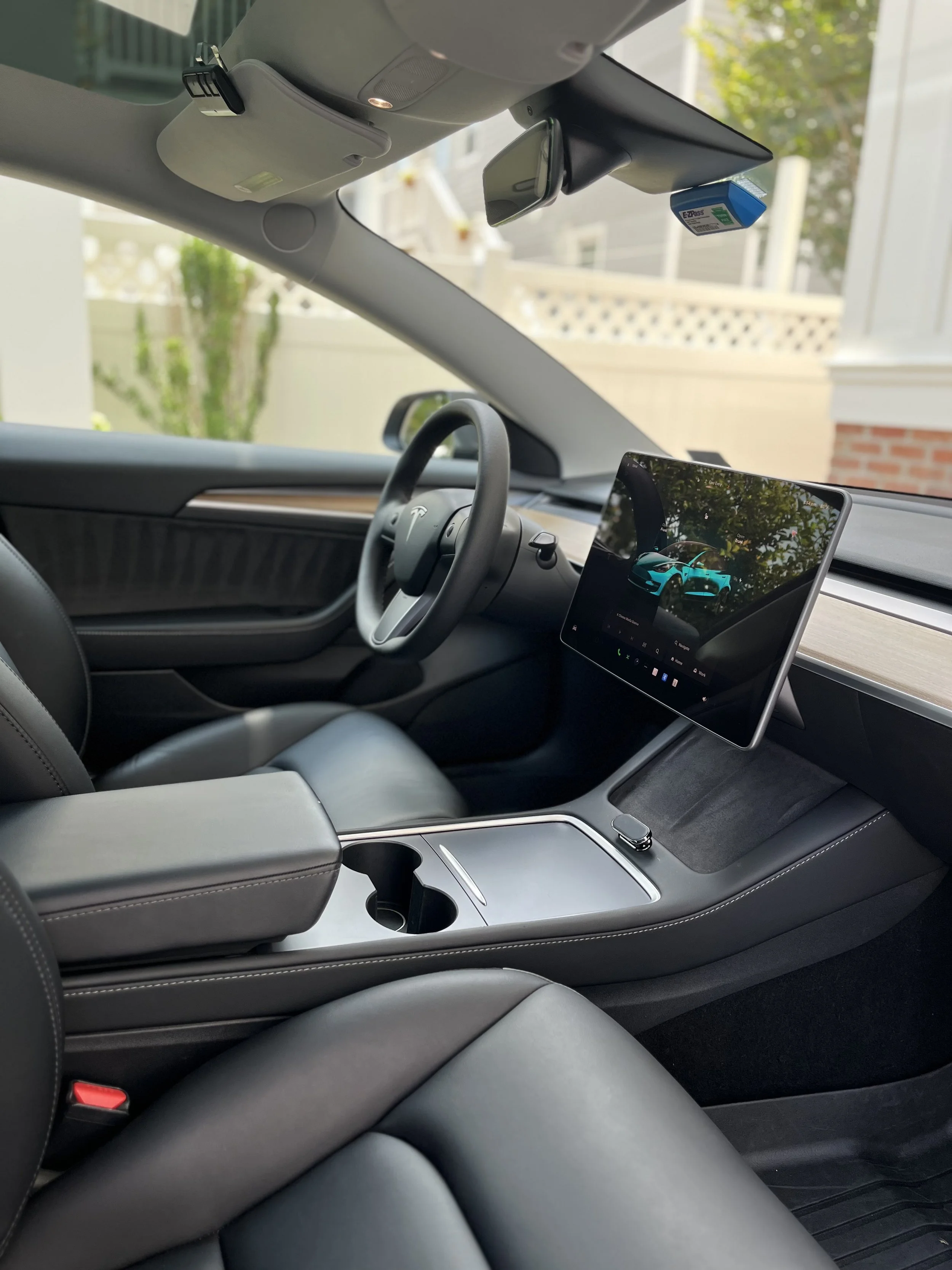 Inside a Tesla vehicle showing the dashboard, steering wheel, central console, touchscreen display, and dashboard camera mount, with a view outside the window of a residential house and a tree.