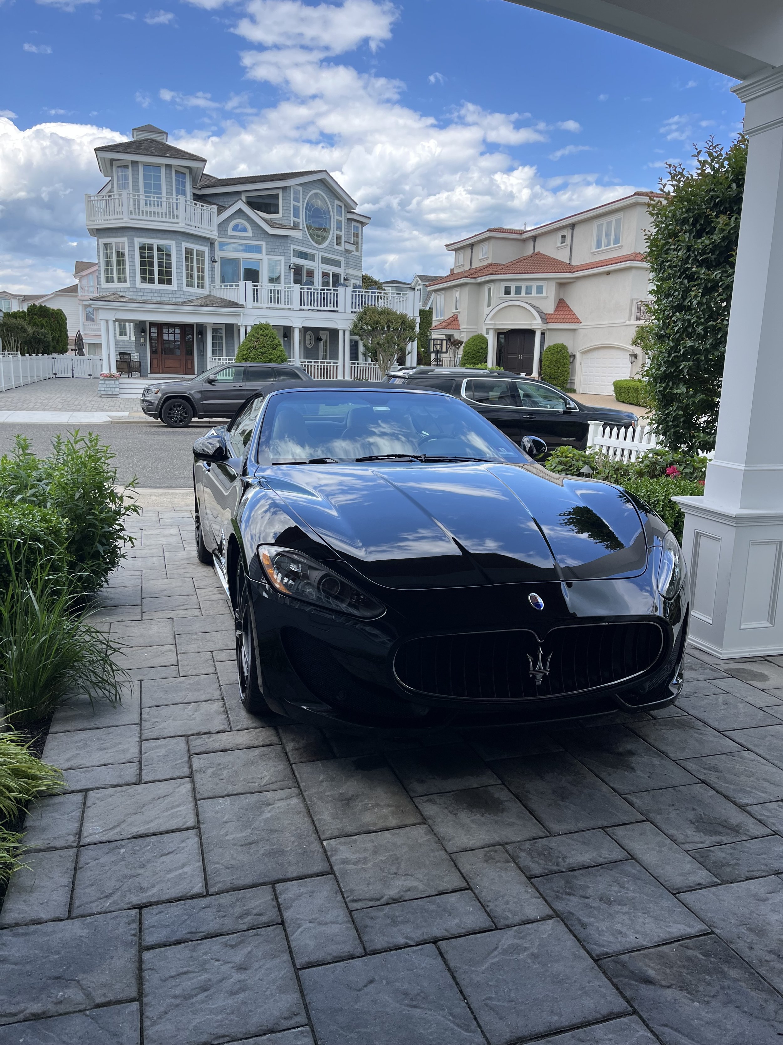 A black Maserati sports car parked on a stone driveway in front of a white house, with other cars and houses visible in the background, under a partly cloudy blue sky.