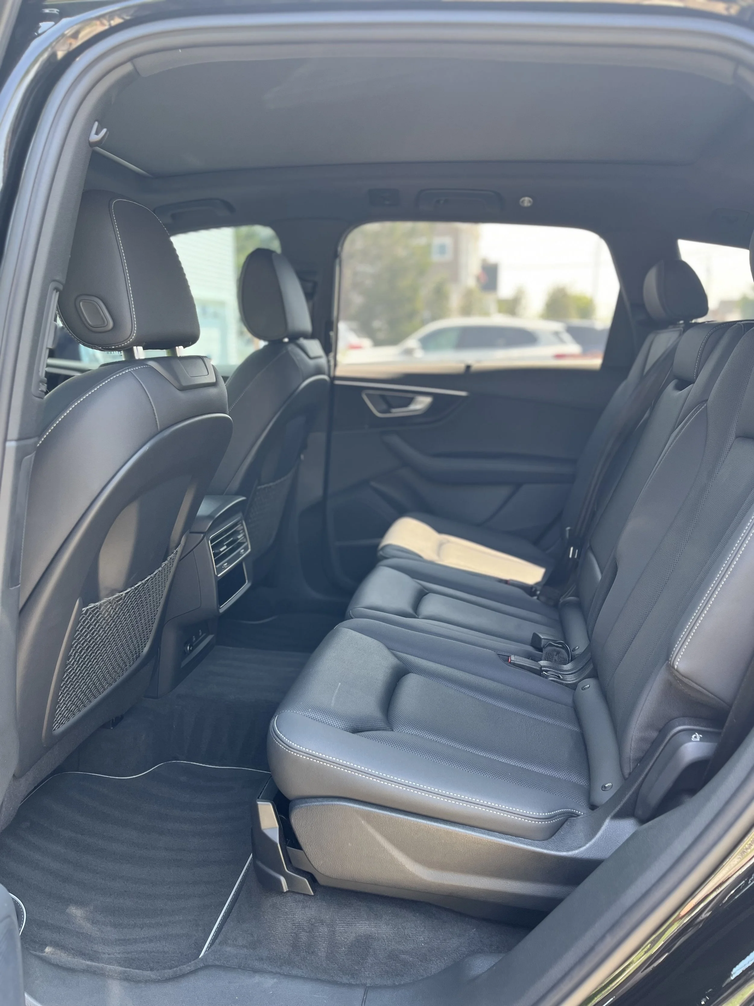 The interior back seat area of a vehicle, showing black seats with white stitching, headrests, a netted storage behind the front seats, and side window views of parked cars outside.