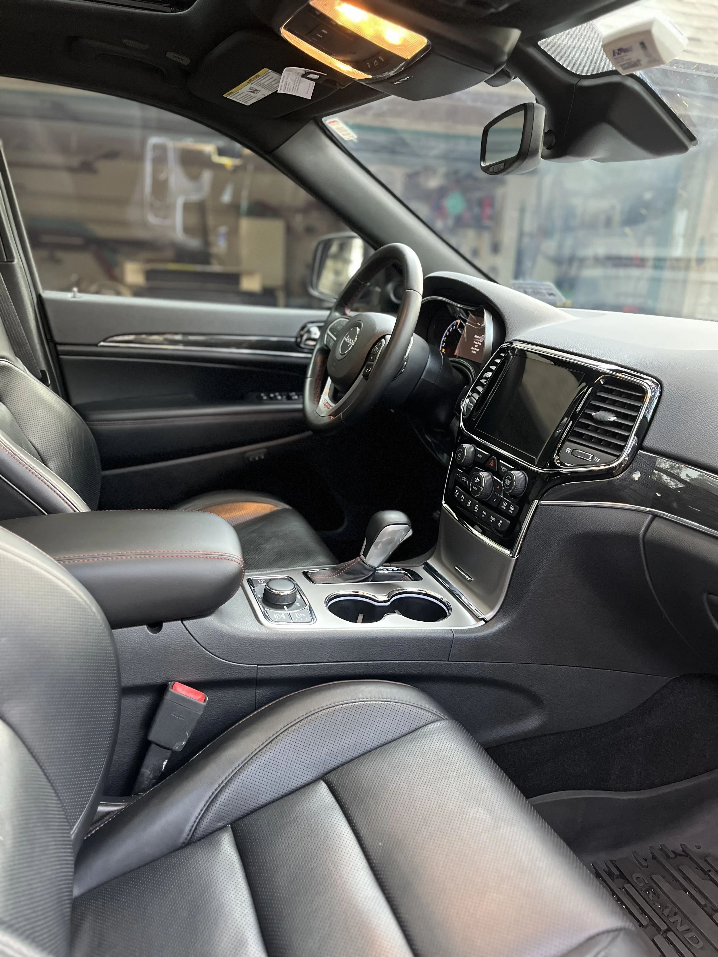 The interior of a modern Jeep vehicle showing the driver's seat, steering wheel, dashboard with touchscreen display, control buttons, gear shift, cup holders, and front passenger seat with black and gray leather upholstery.