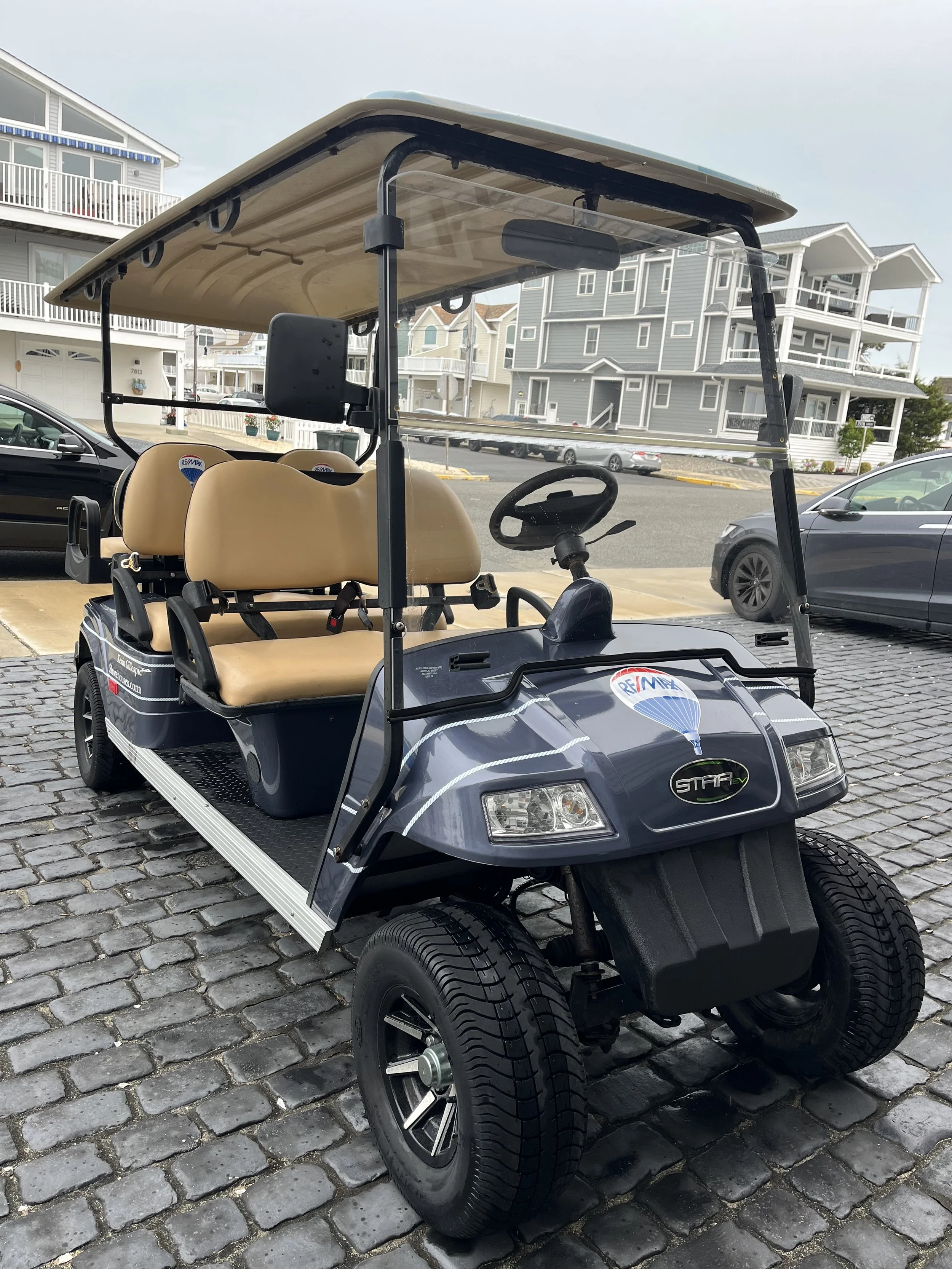 A black golf cart with tan seats parked on a cobblestone street in front of residential buildings.
