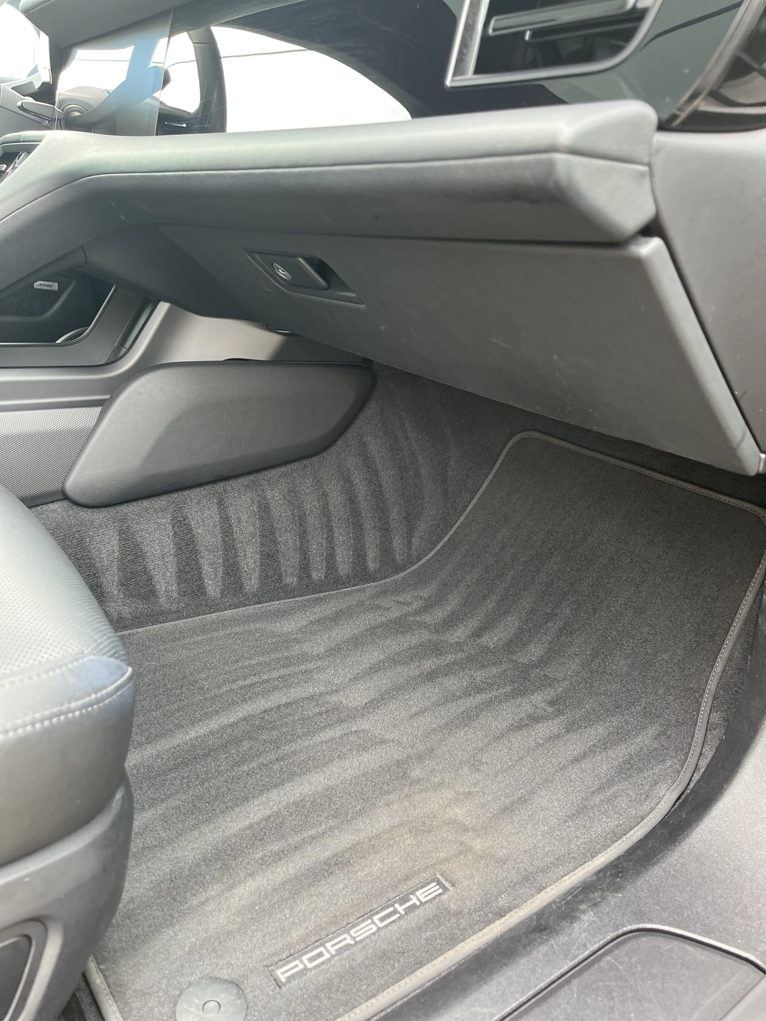 Interior floor of a Porsche vehicle showing a black carpeted floor with the Porsche logo on the floor mat, part of a seat, and part of the center console.