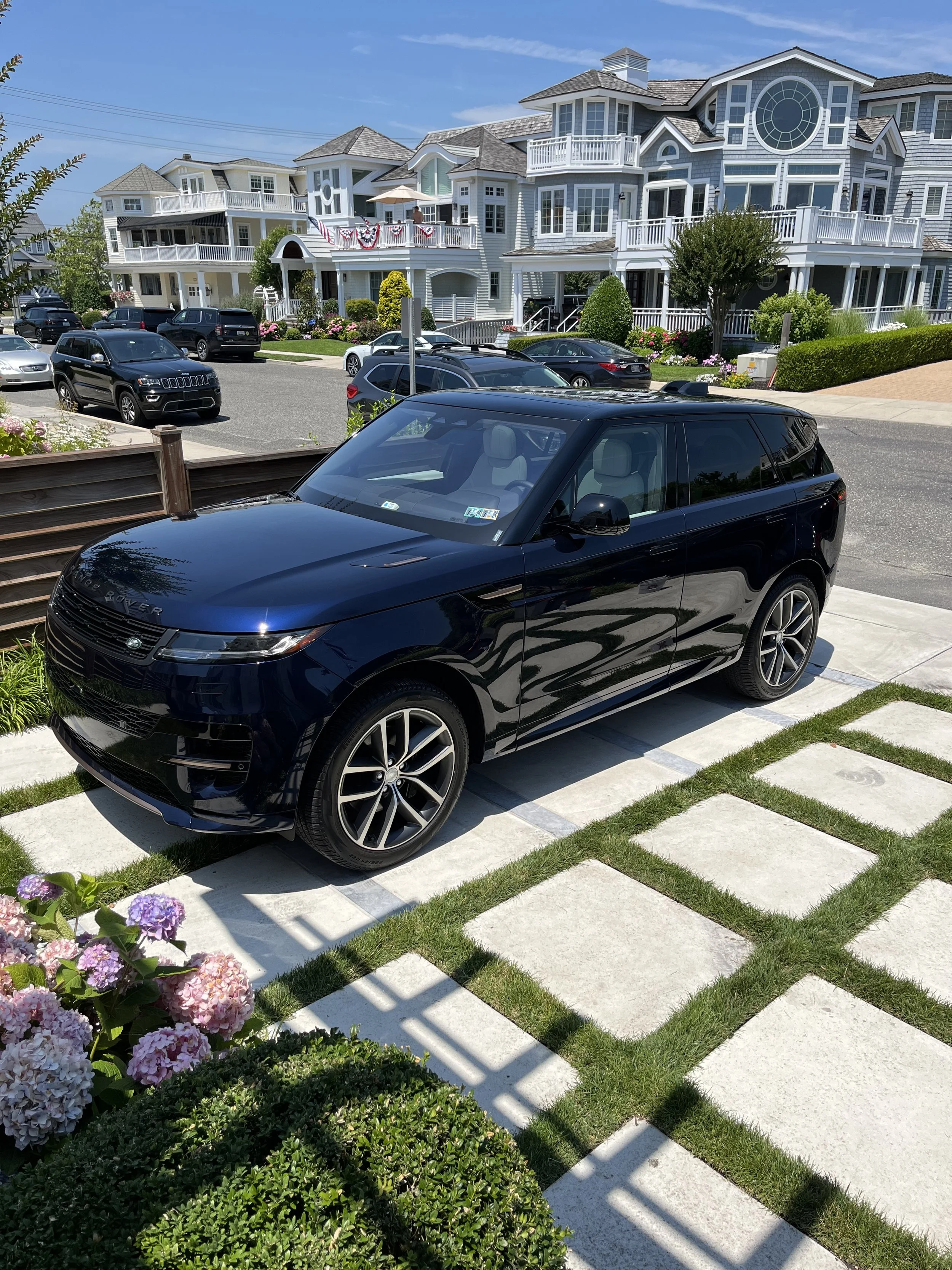 A black Range Rover SUV parked on a driveway with a modern, multi-story house in the background. The house has blue siding, white trim, multiple balconies, and a large circular window. There are colorful flowers and green shrubs in the yard, with blu