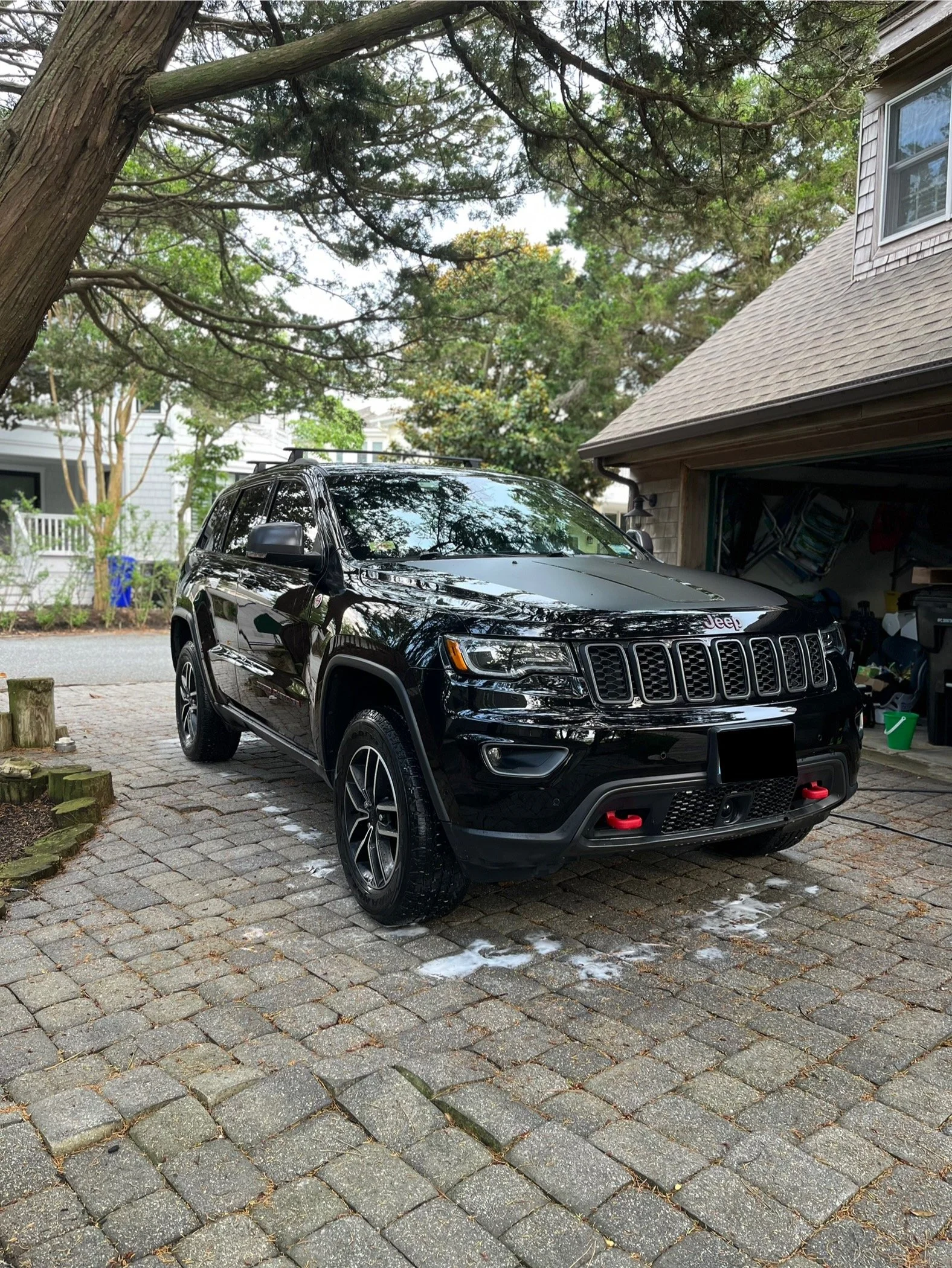 A black Jeep SUV parked in a driveway, with soap suds on the cobblestone ground around it, in front of a house with a garage and trees overhead.