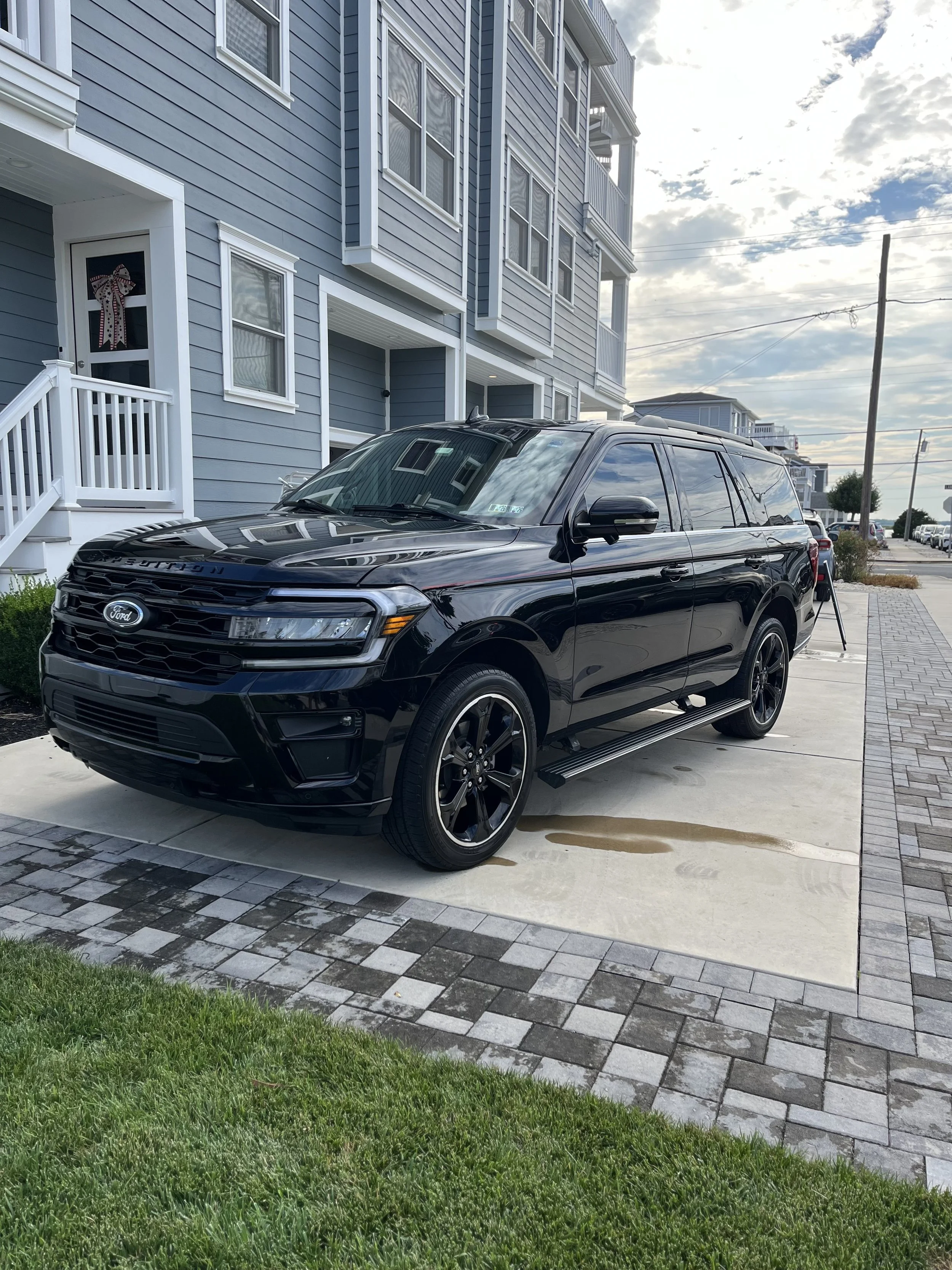 A black Ford Expedition SUV parked on a concrete driveway in front of a blue multi-story residential building with white trim and balconies. There are multiple windows, some with window coverings, and a door with decorative ribbon embellishment. The 