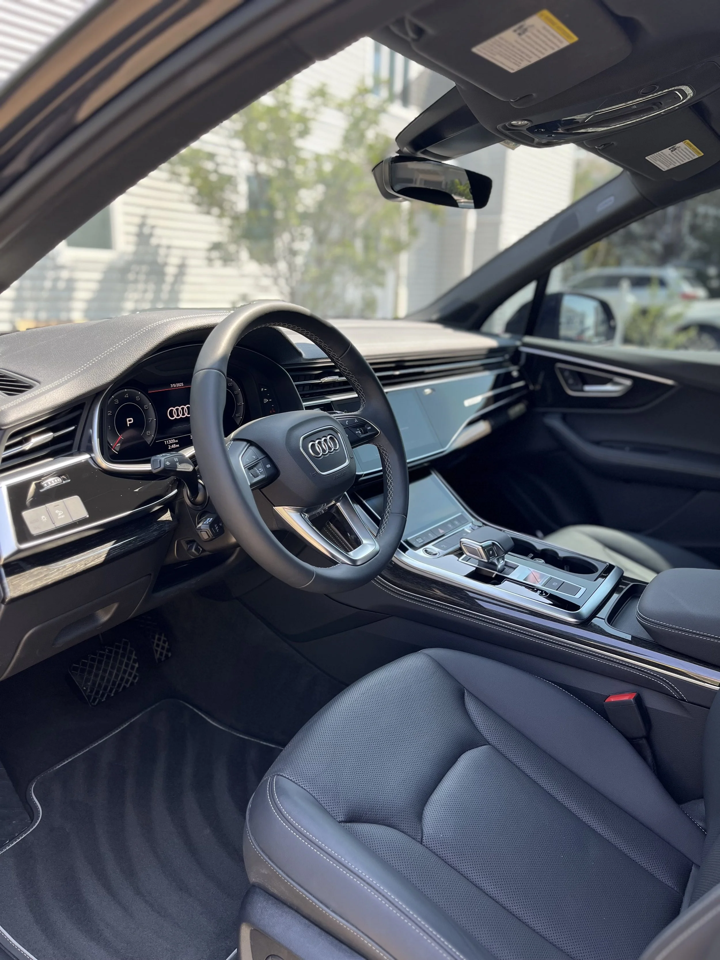Interior of a modern luxury vehicle, showing the driver's seat, steering wheel with Audi logo, digital dashboard, center console with gear shifter, and rearview mirror, with a view of outdoors through windows.