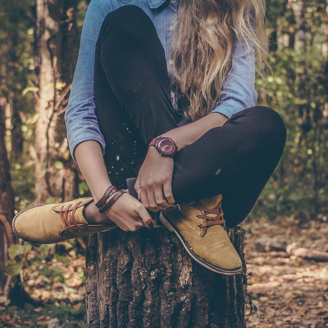 Person sitting on a tree stump in the woods, wearing a denim shirt, black pants, tan boots, and a red wristwatch.