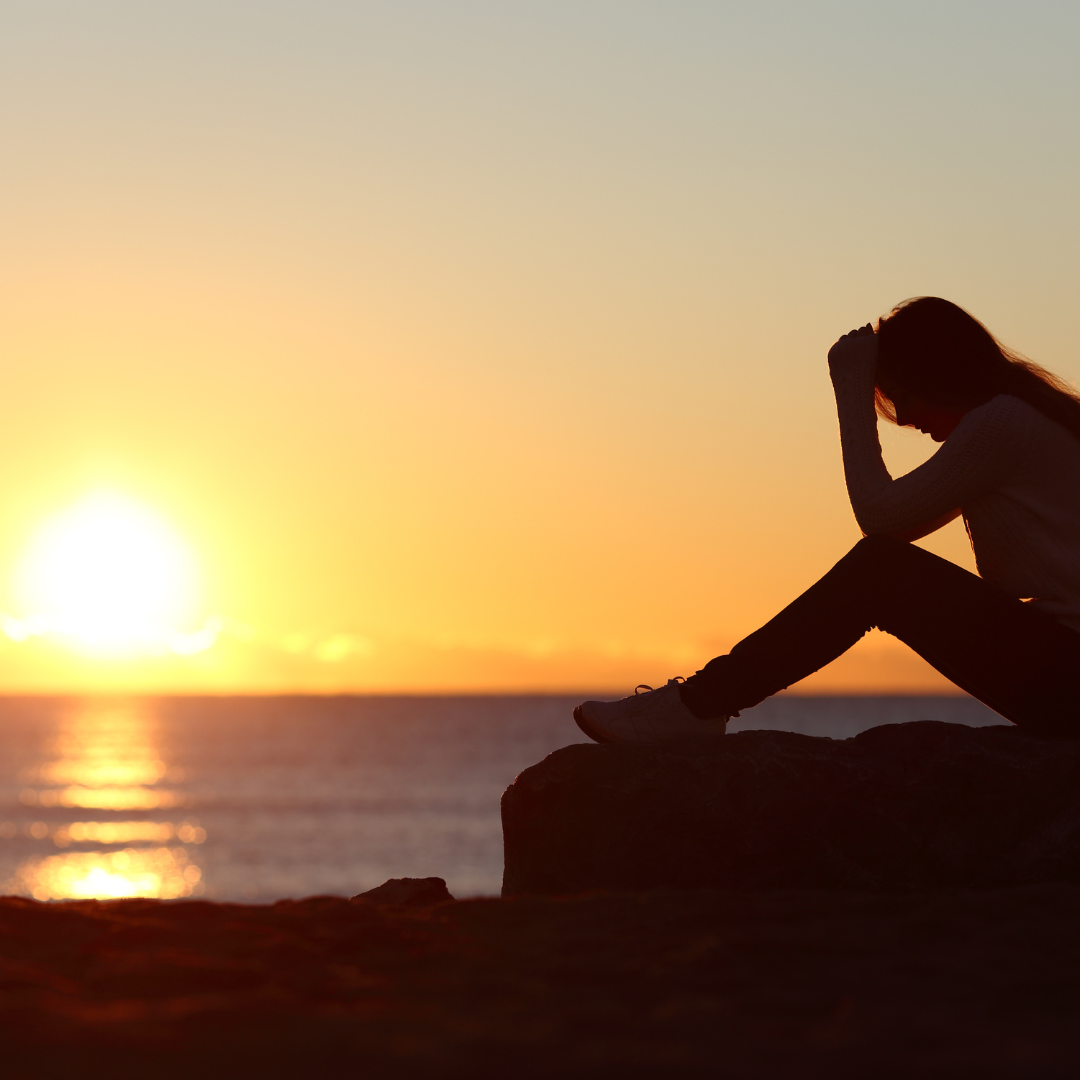 Silhouette of a woman sitting on a rock at sunset by the ocean, with her head in her hand.