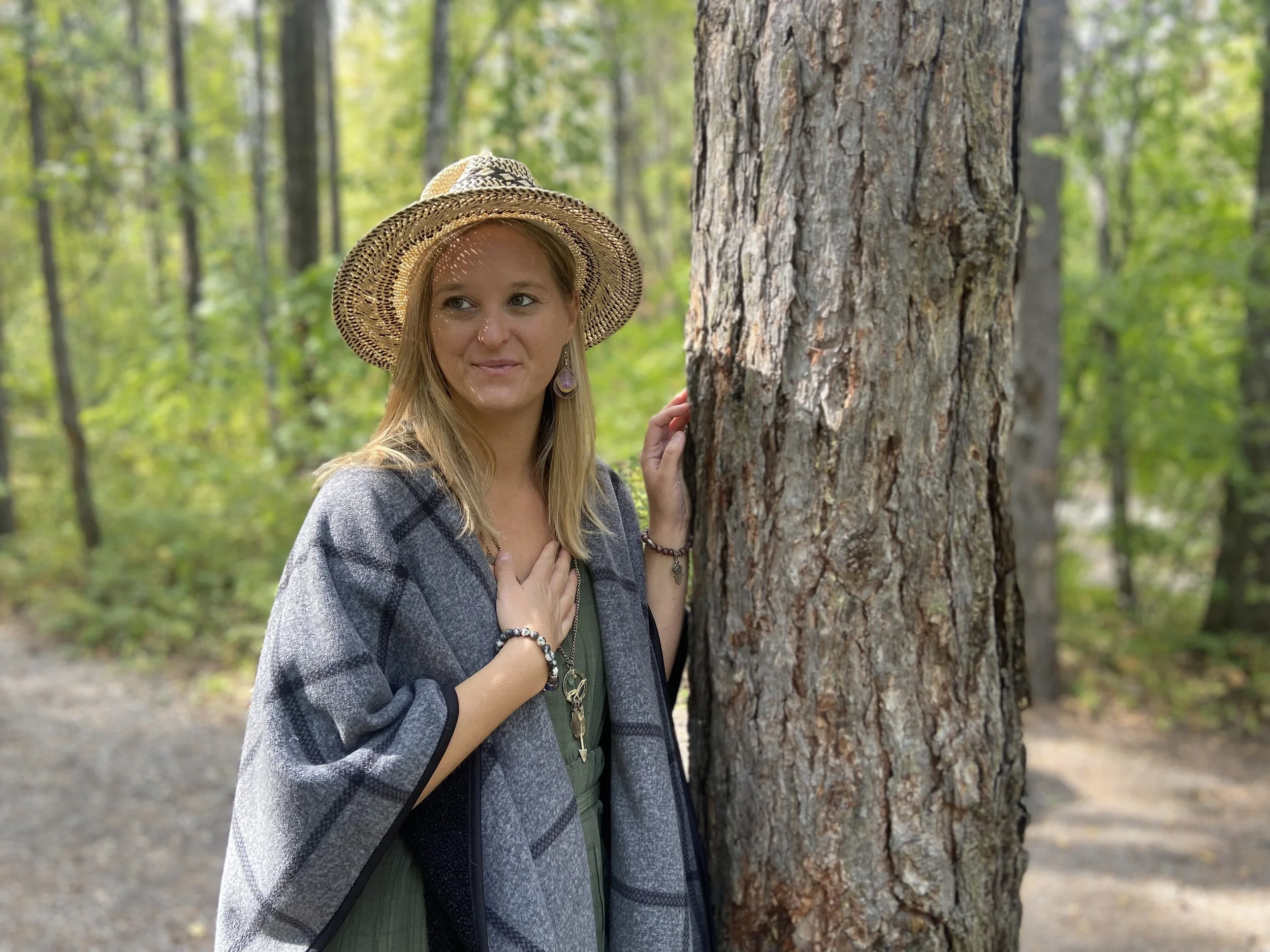 A woman wearing a wide-brimmed straw hat and jewelry stands next to a large tree in a forest with green foliage.