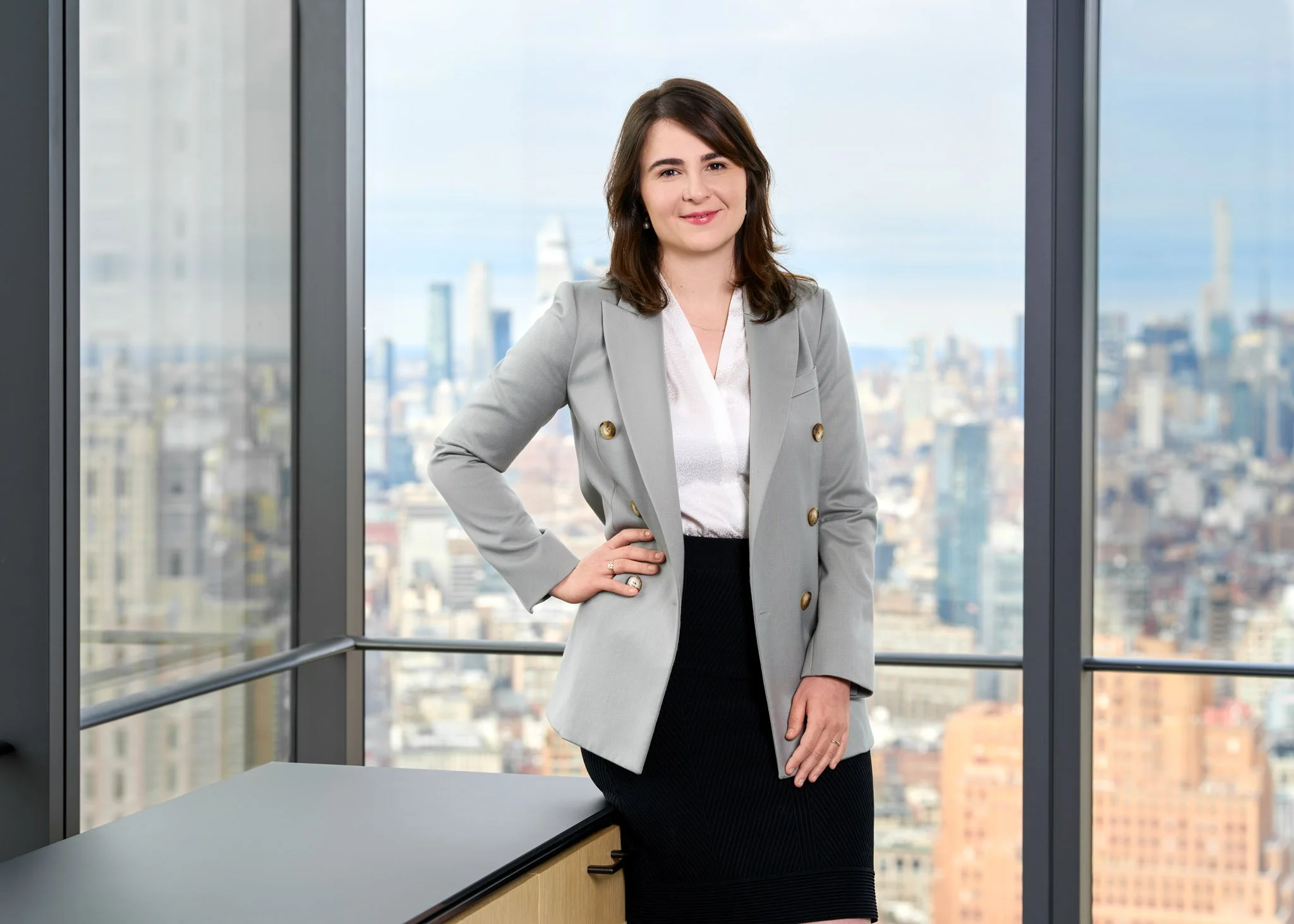 Executive headshot of a woman in a light gray blazer standing confidently by the window with city views. A polished, professional corporate portrait in natural light.
