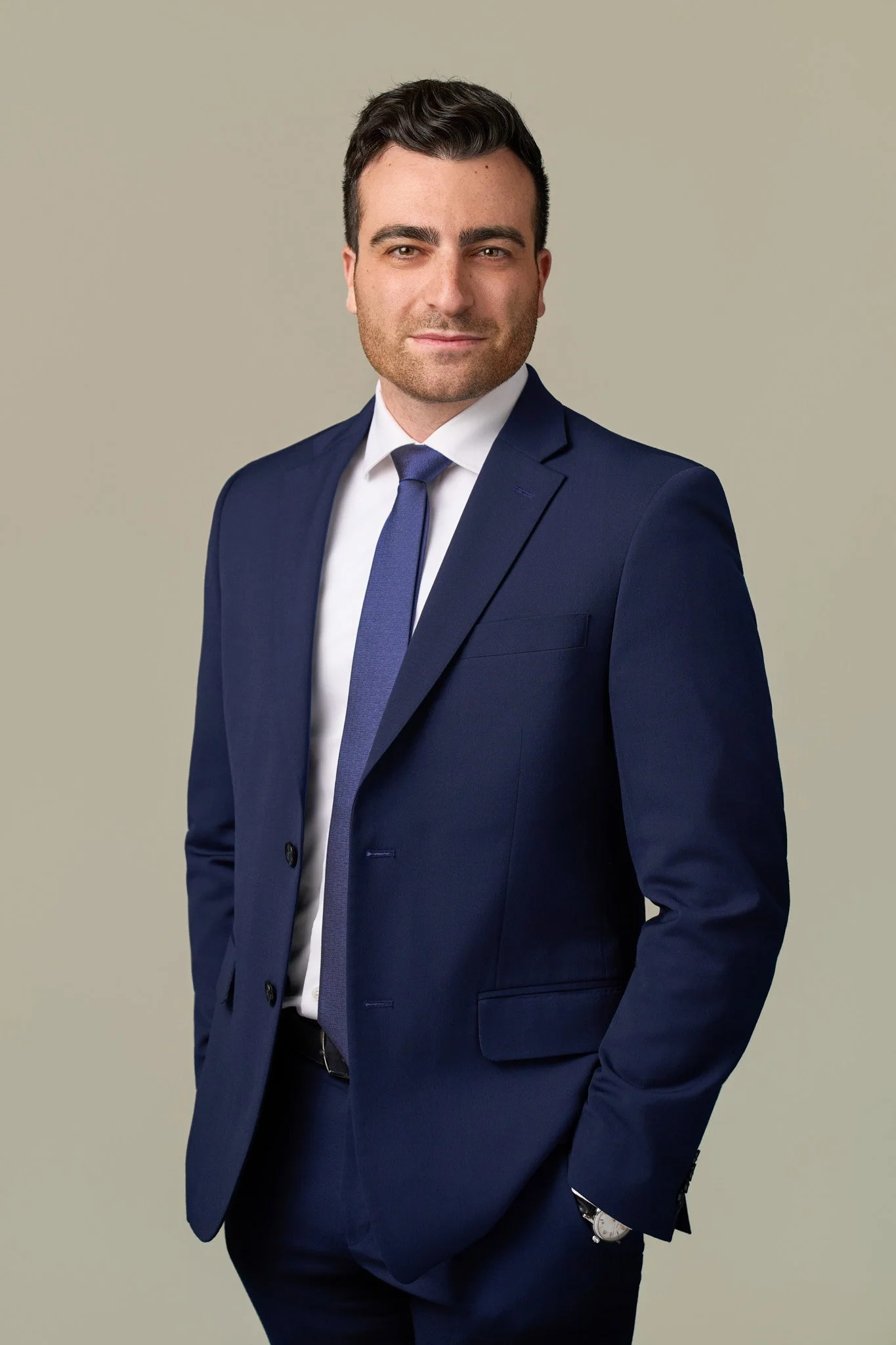 Male executive in a navy suit and light blue tie with hands in pockets, NYC corporate portrait on neutral studio backdrop