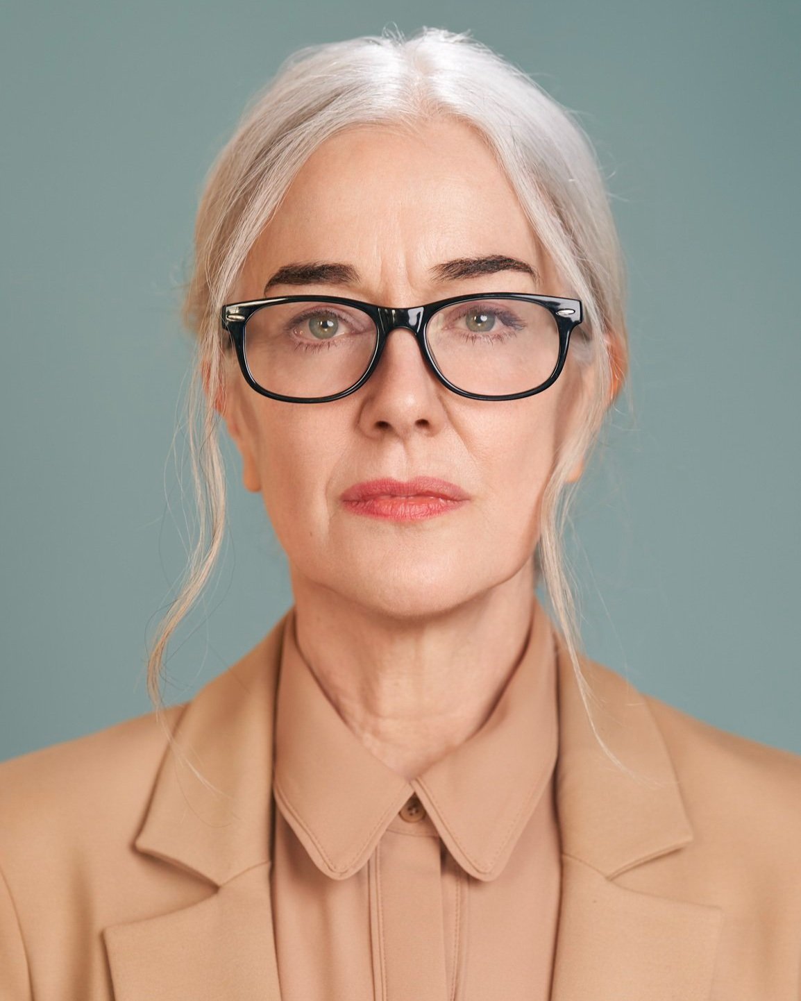Actor headshot of a woman with silver hair and glasses, composed and serious expression, wearing a neutral-toned jacket, photographed against a soft green studio background in NYC by David Pexton.