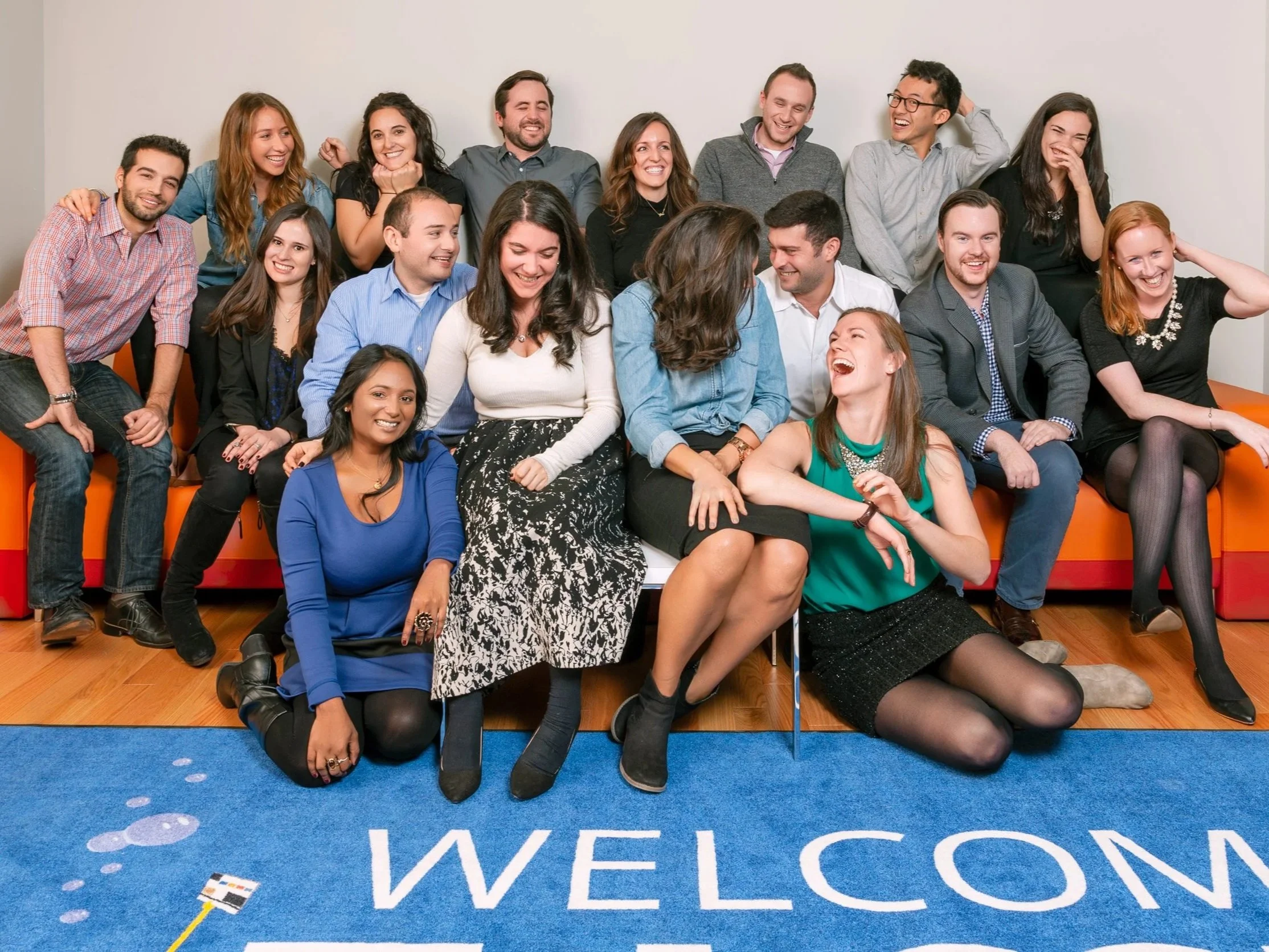 Casual company team portrait of sixteen people on an orange sofa and blue welcome rug, corporate group photography in New York City
