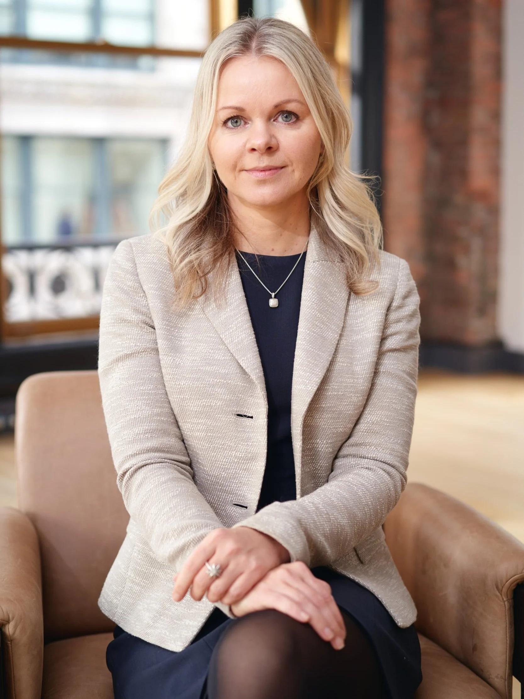 Female executive in a cream tweed blazer seated against an exposed brick wall, professional headshot photographed in a Manhattan office
