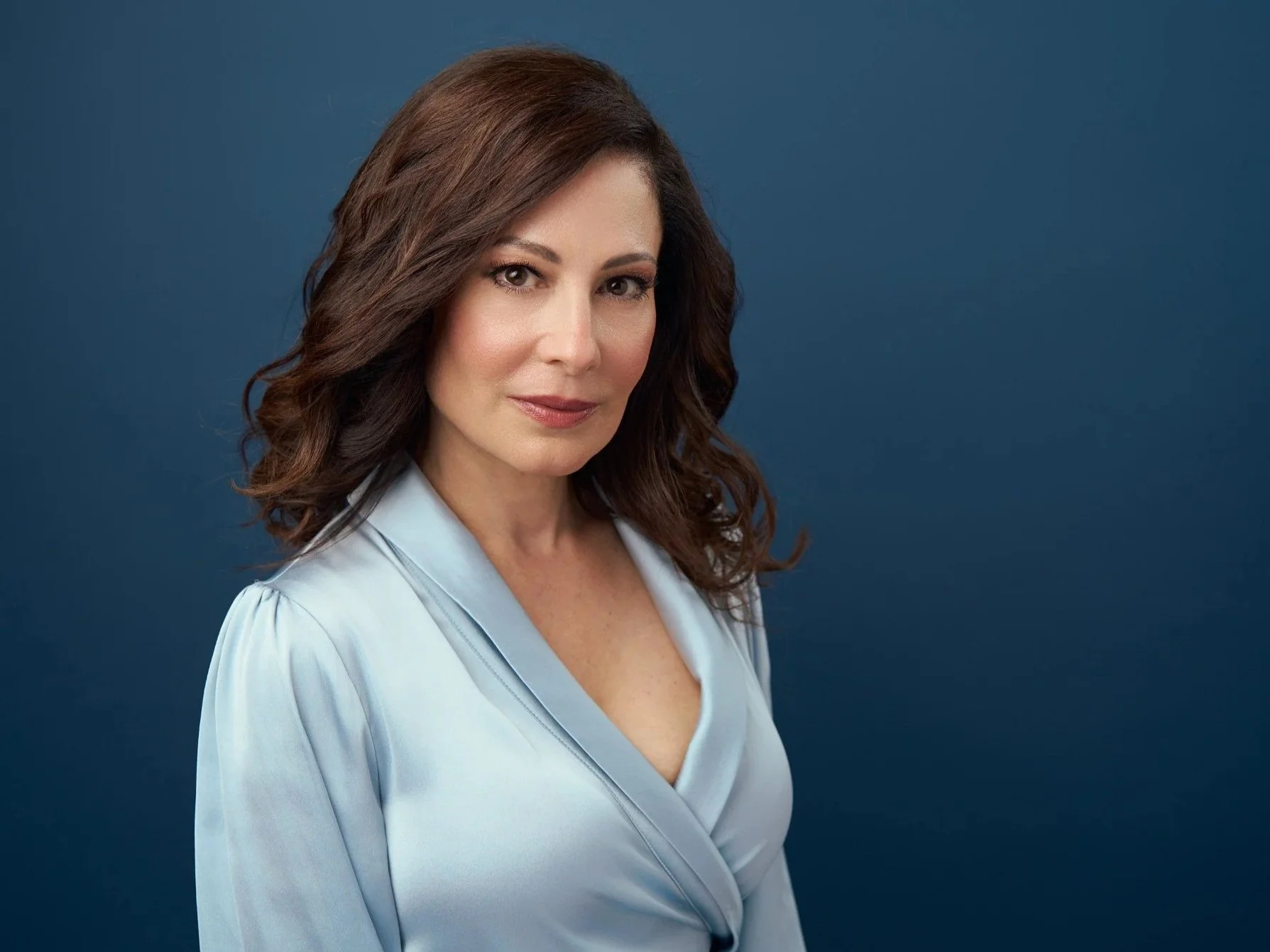 A woman with shoulder-length wavy brown hair and fair skin wearing a light blue blouse, posing against a dark blue background.