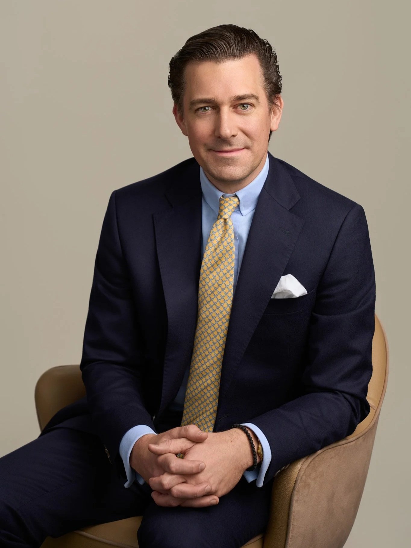 Male executive in a navy suit and gold tie seated with hands clasped, corporate studio headshot photographed in New York City