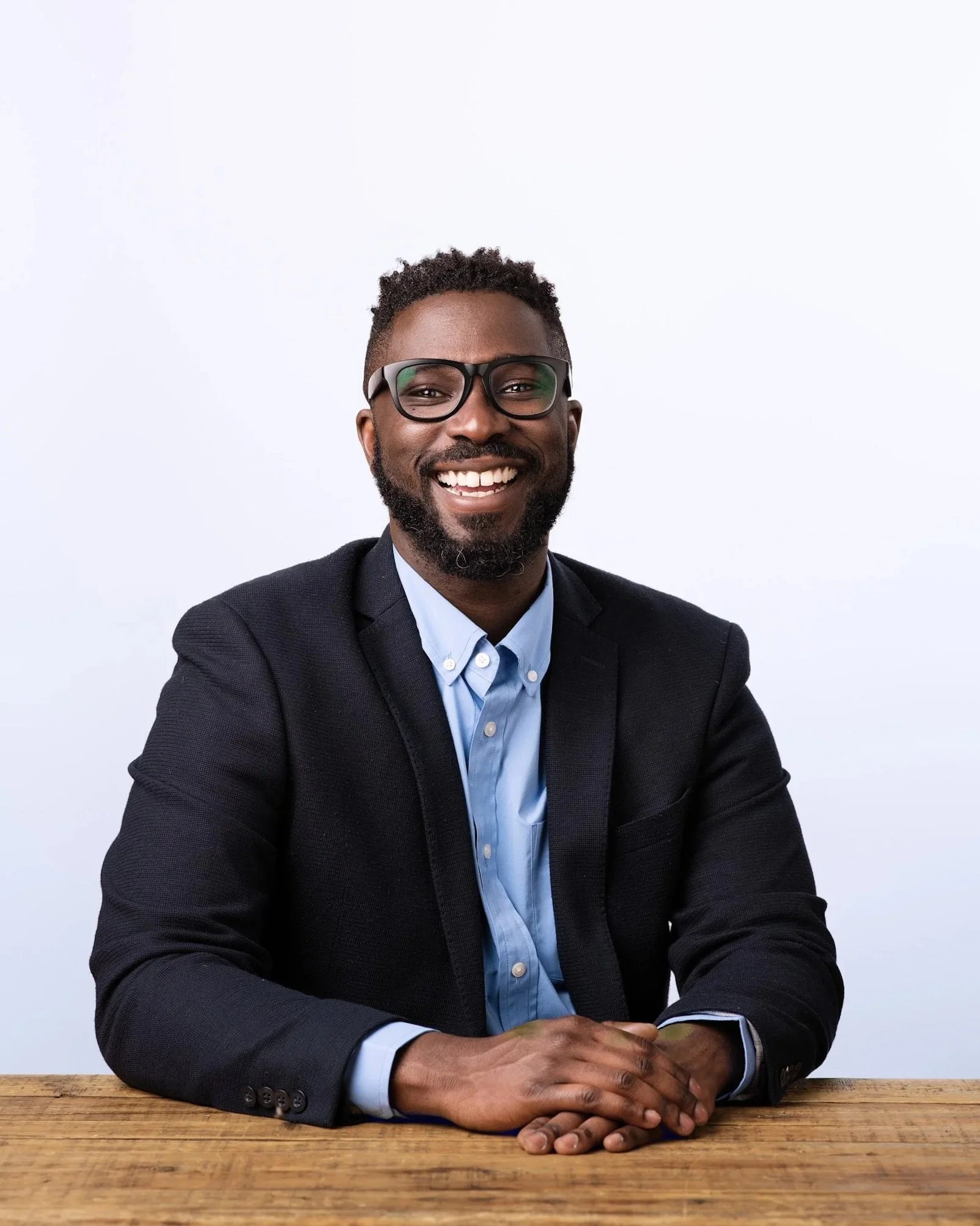 A smiling man with glasses wearing a suit and blue shirt, sitting at a wooden table against a plain white wall.