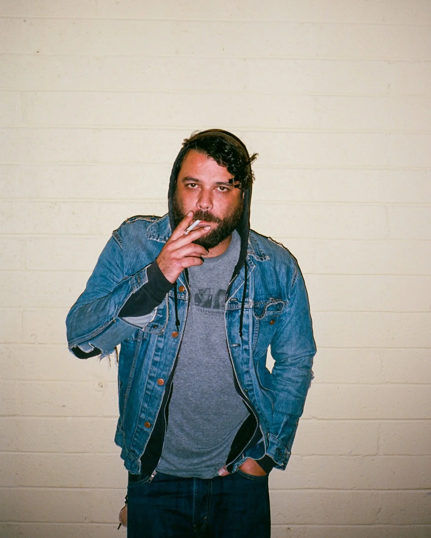 Editorial portrait of a man in a denim jacket and hoodie standing against a painted brick wall, direct flash lighting and casual urban style, photographed in New York City by David Pexton.