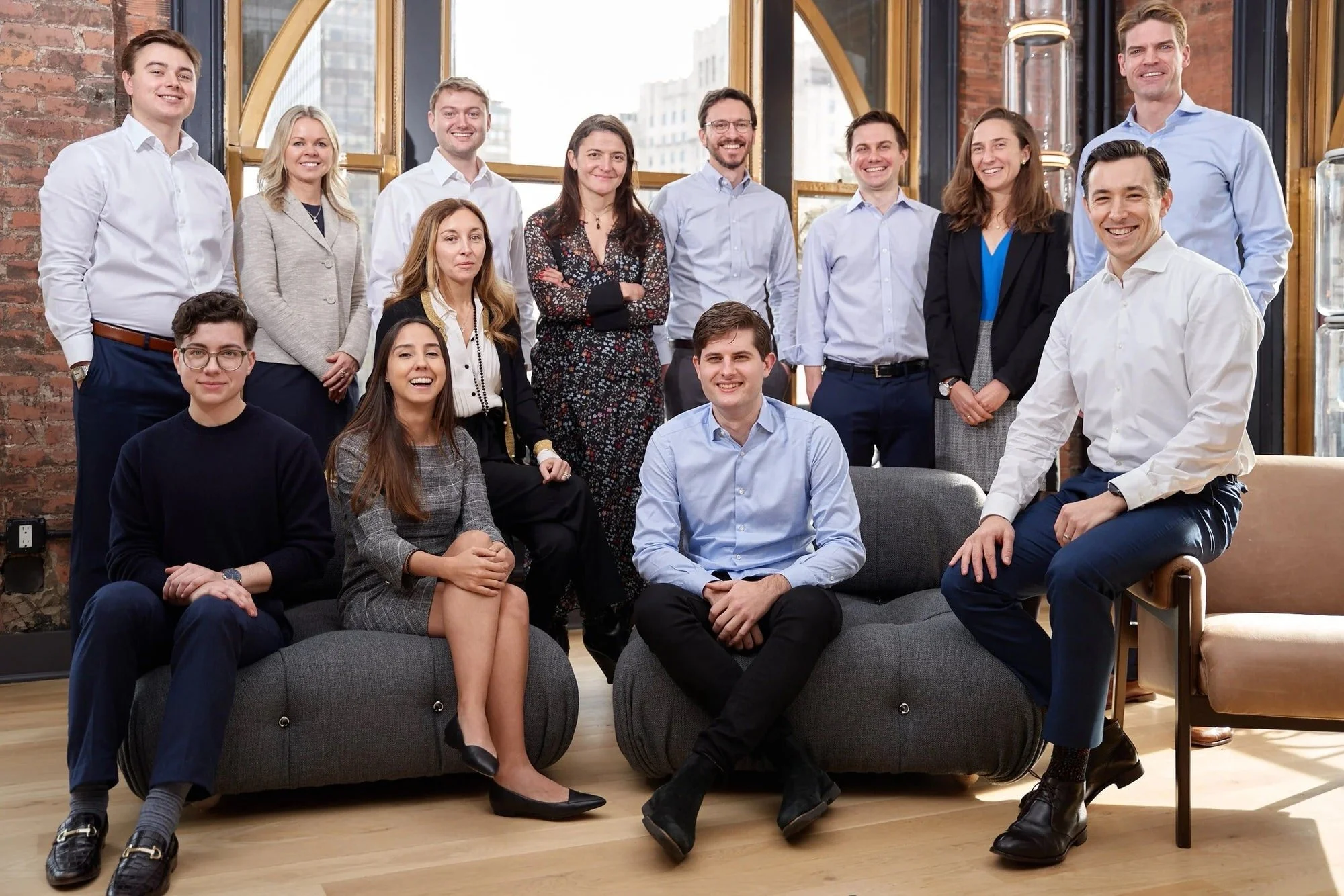 Corporate team portrait of thirteen professionals on grey sofas in a NYC office with exposed brick and tall arched windows, photographed by David Pexton