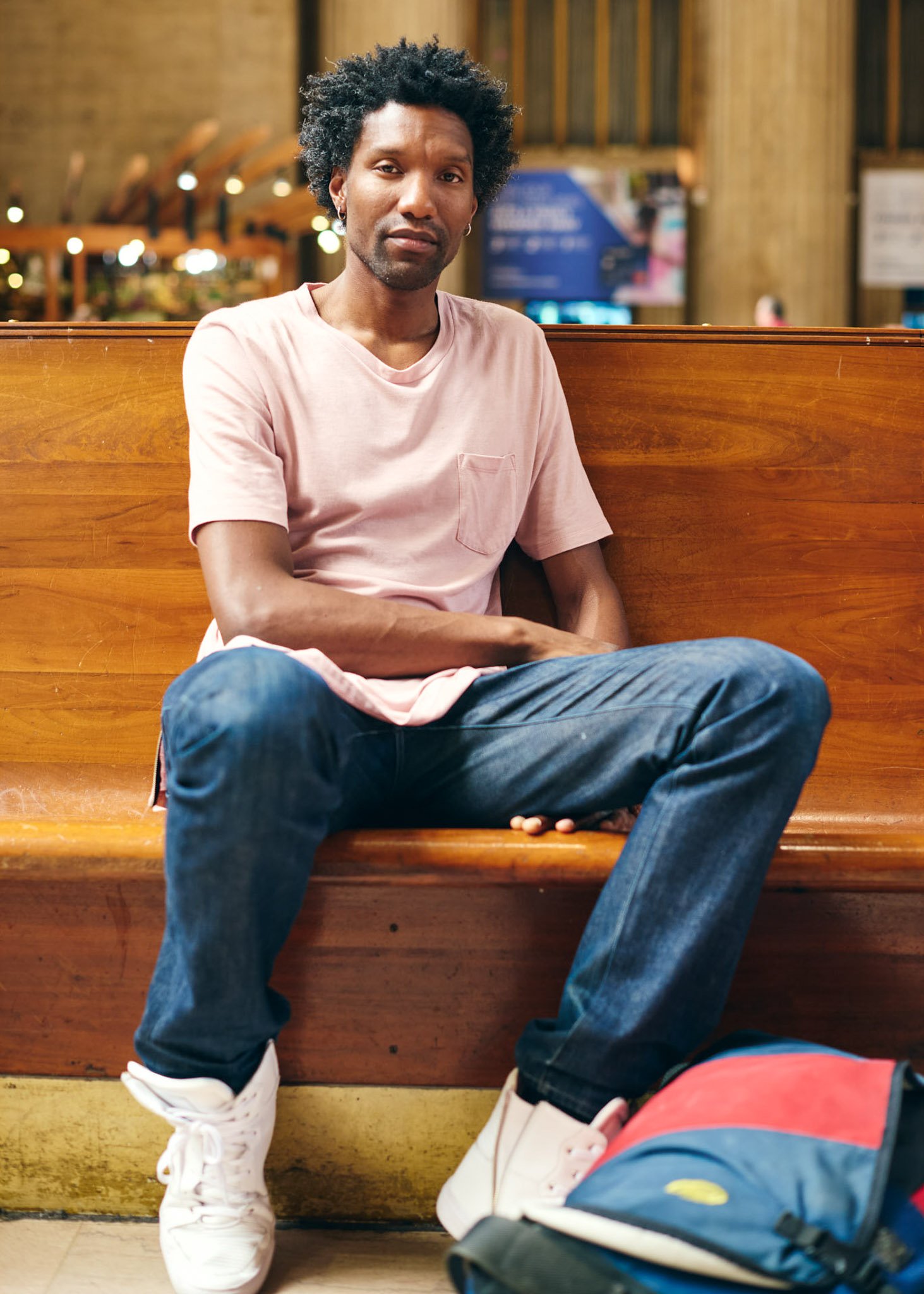 Lifestyle portrait of a man sitting on a wooden bench inside an Amtrak station, wearing a light pink T-shirt and jeans, photographed for National Magazine by David Pexton Photography.