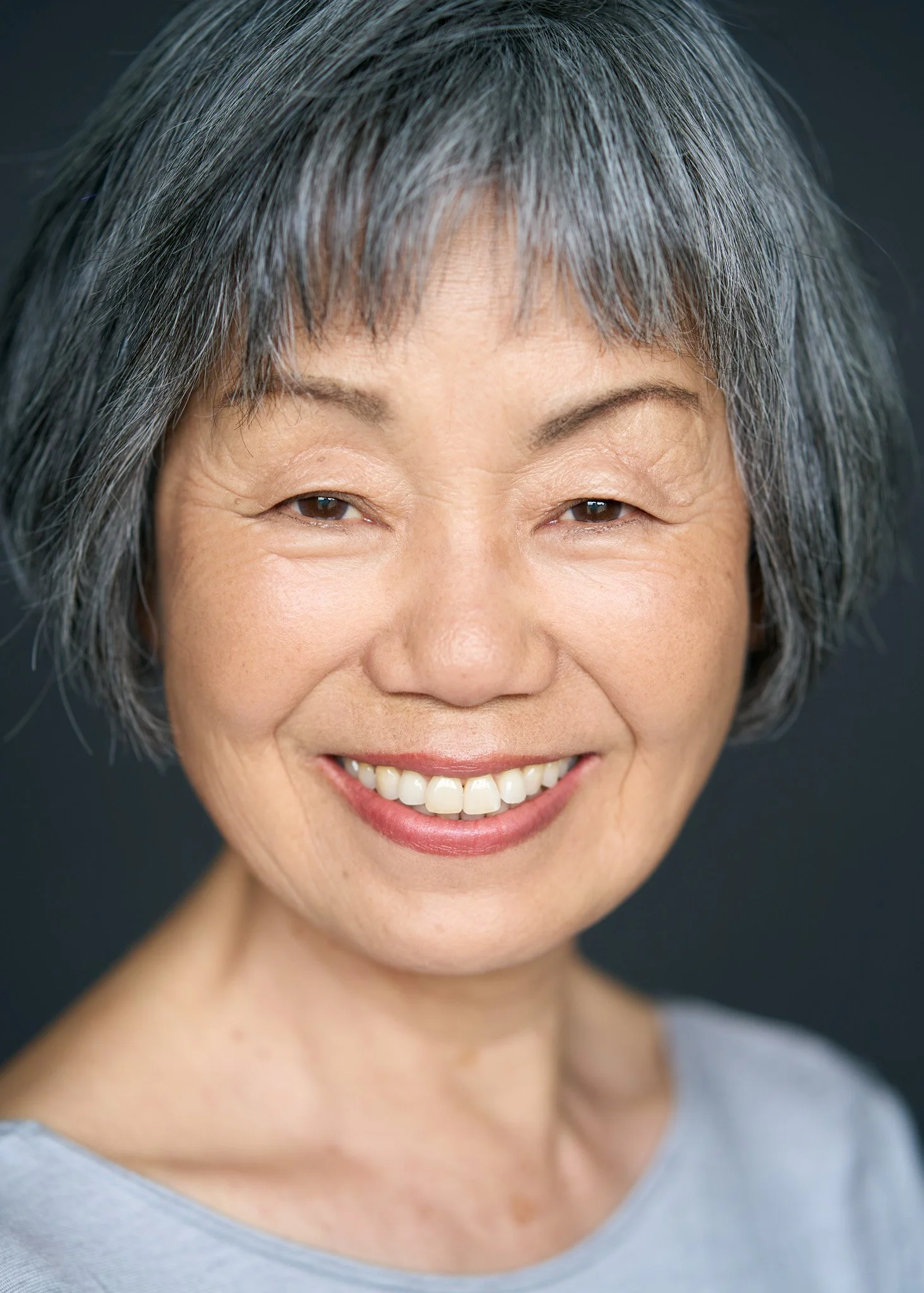 Actor headshot of a mature woman with short gray hair smiling against a soft blue studio background, clean natural lighting and shallow depth of field, photographed in New York City by David Pexton.