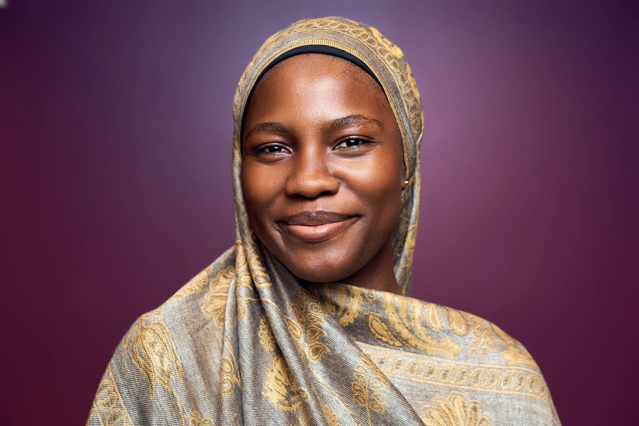 Portrait of a woman wearing a patterned headscarf with a purple background.
