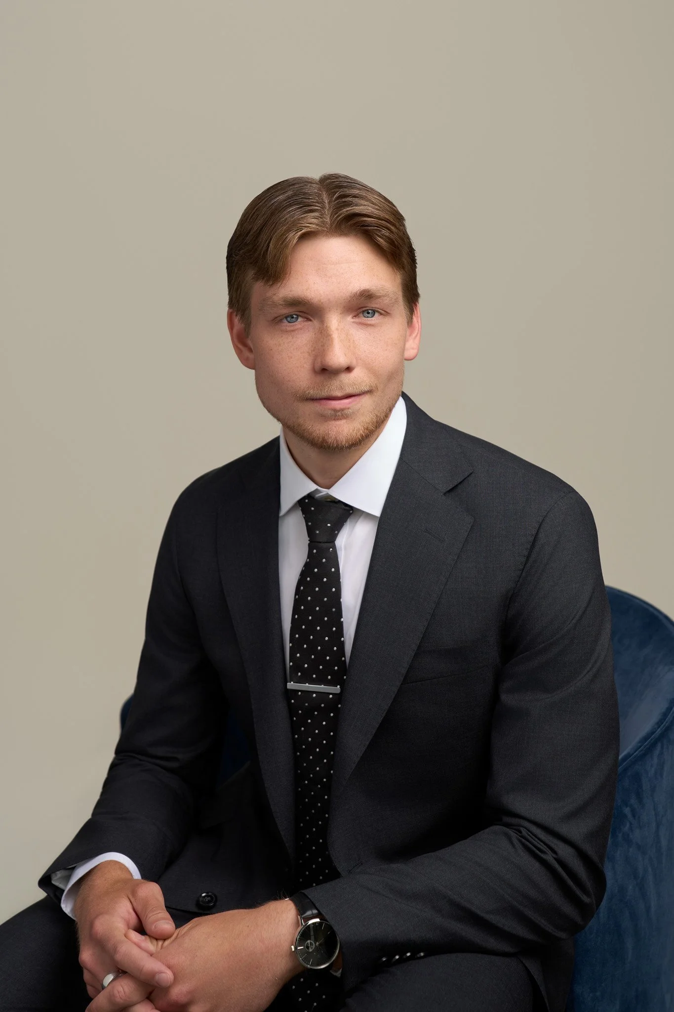 Young professional man in a black suit and polka-dot tie seated on blue armchair, NYC executive headshot on neutral backdrop