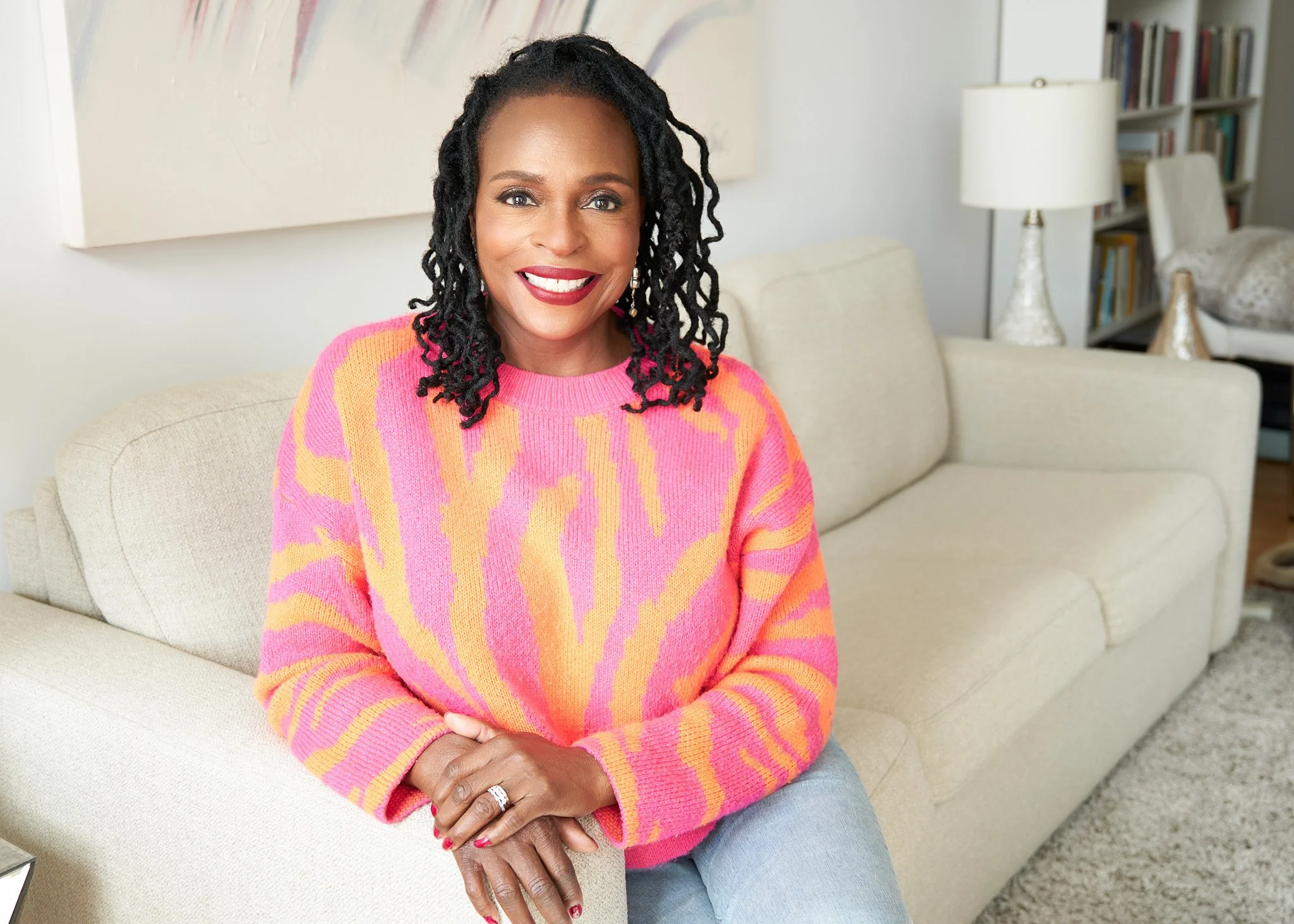 Smiling woman seated on a light sofa in a bright NYC living room, wearing a pink and orange sweater, hands resting in her lap, with bookshelves and a lamp in the background