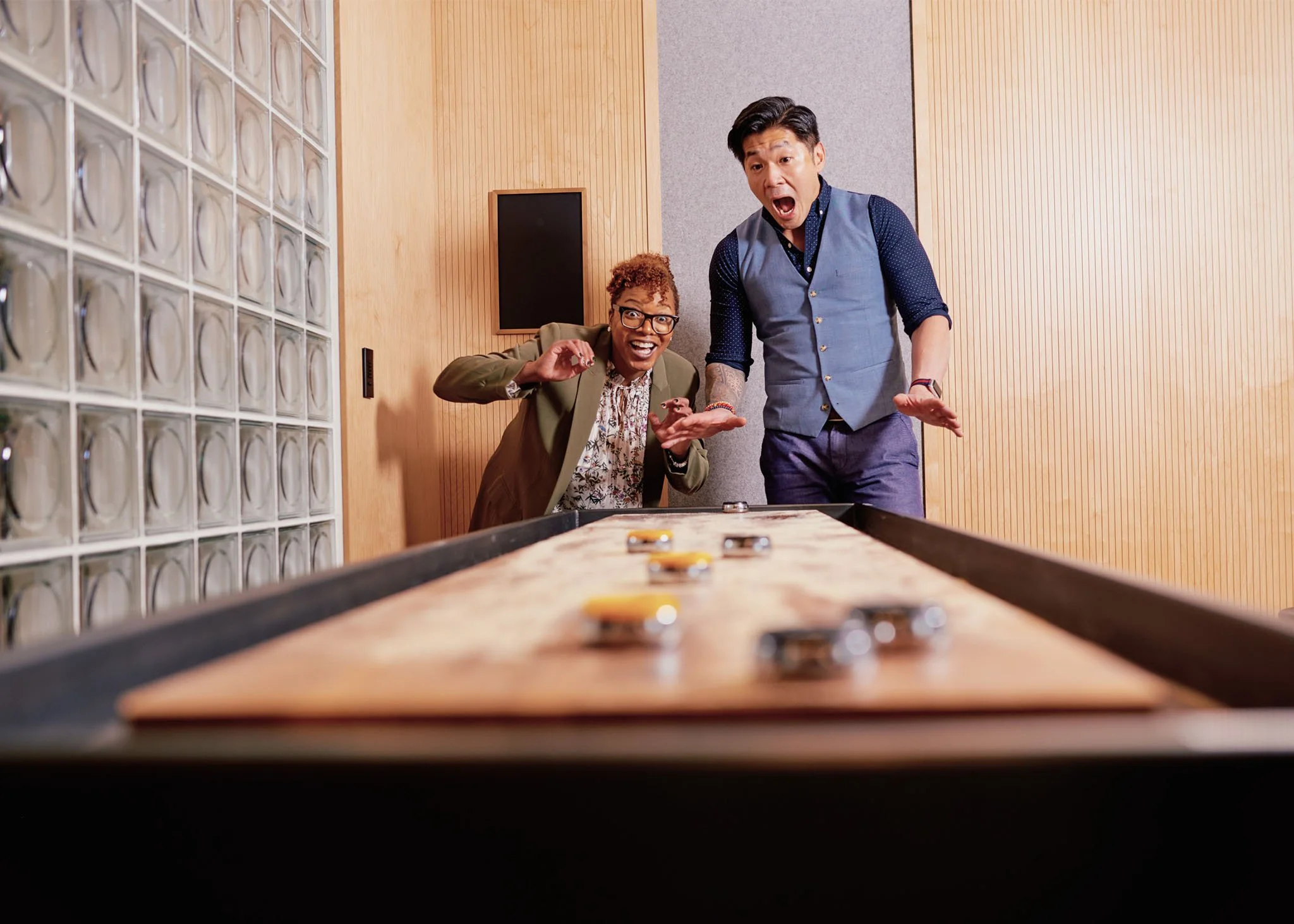 Corporate lifestyle photograph of two colleagues playing shuffleboard in a New York City office, reacting with surprise and laughter
