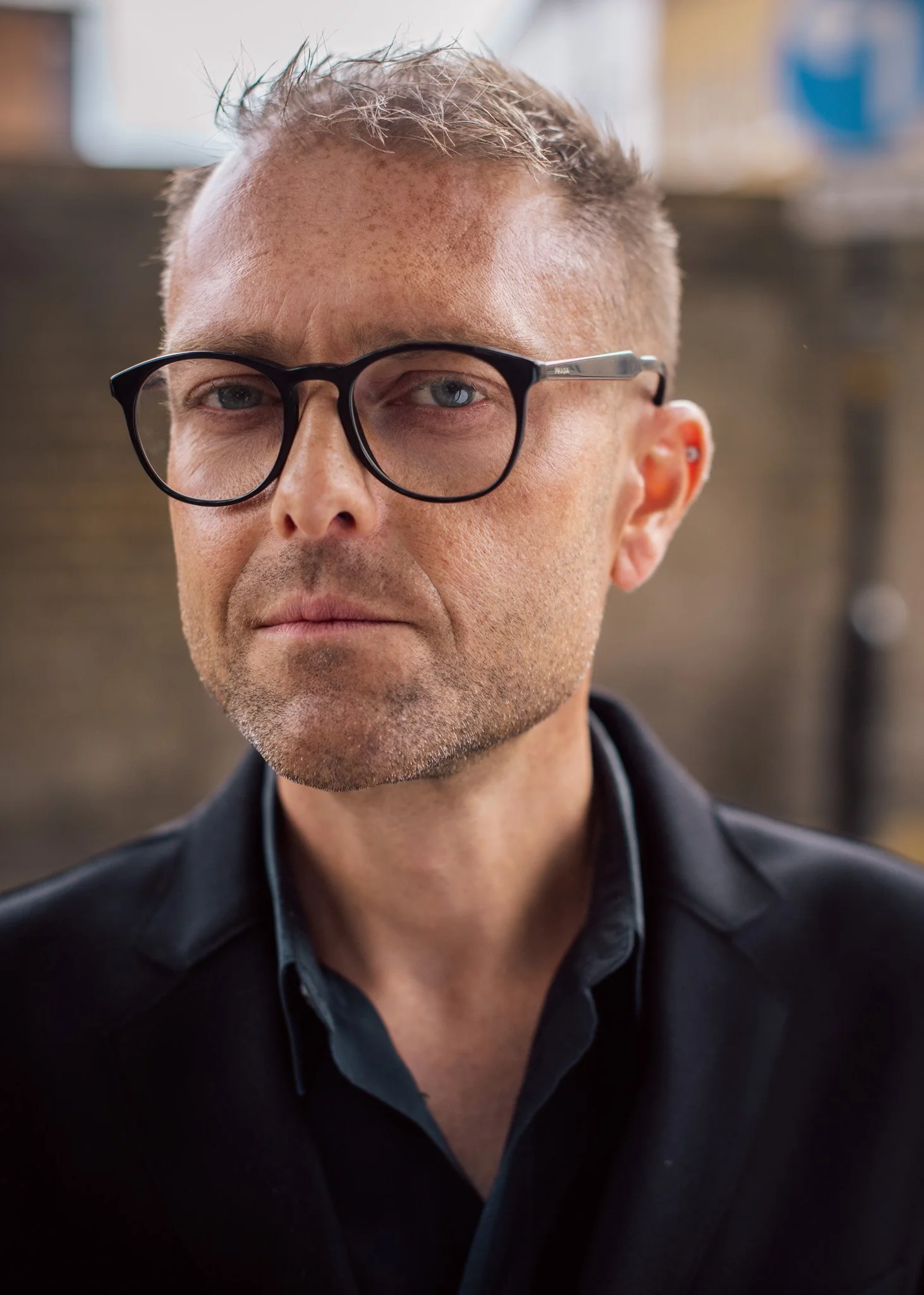 Close-up image of a middle-aged man with short, light-colored hair, glasses, wearing a black shirt and jacket, looking serious against an urban background.