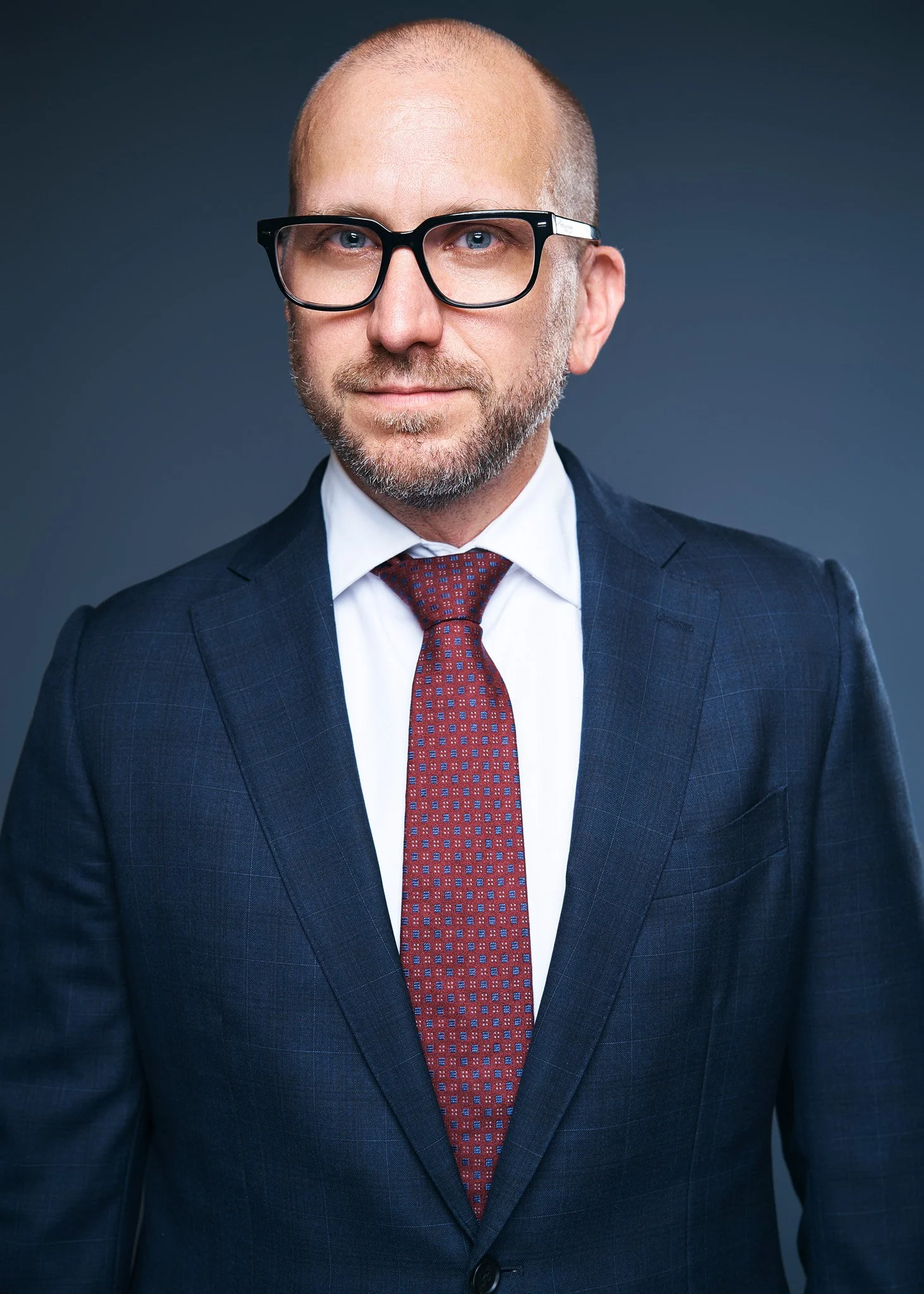 Male executive in a navy suit and burgundy tie on a dark blue background, professional corporate headshot photographed in NYC